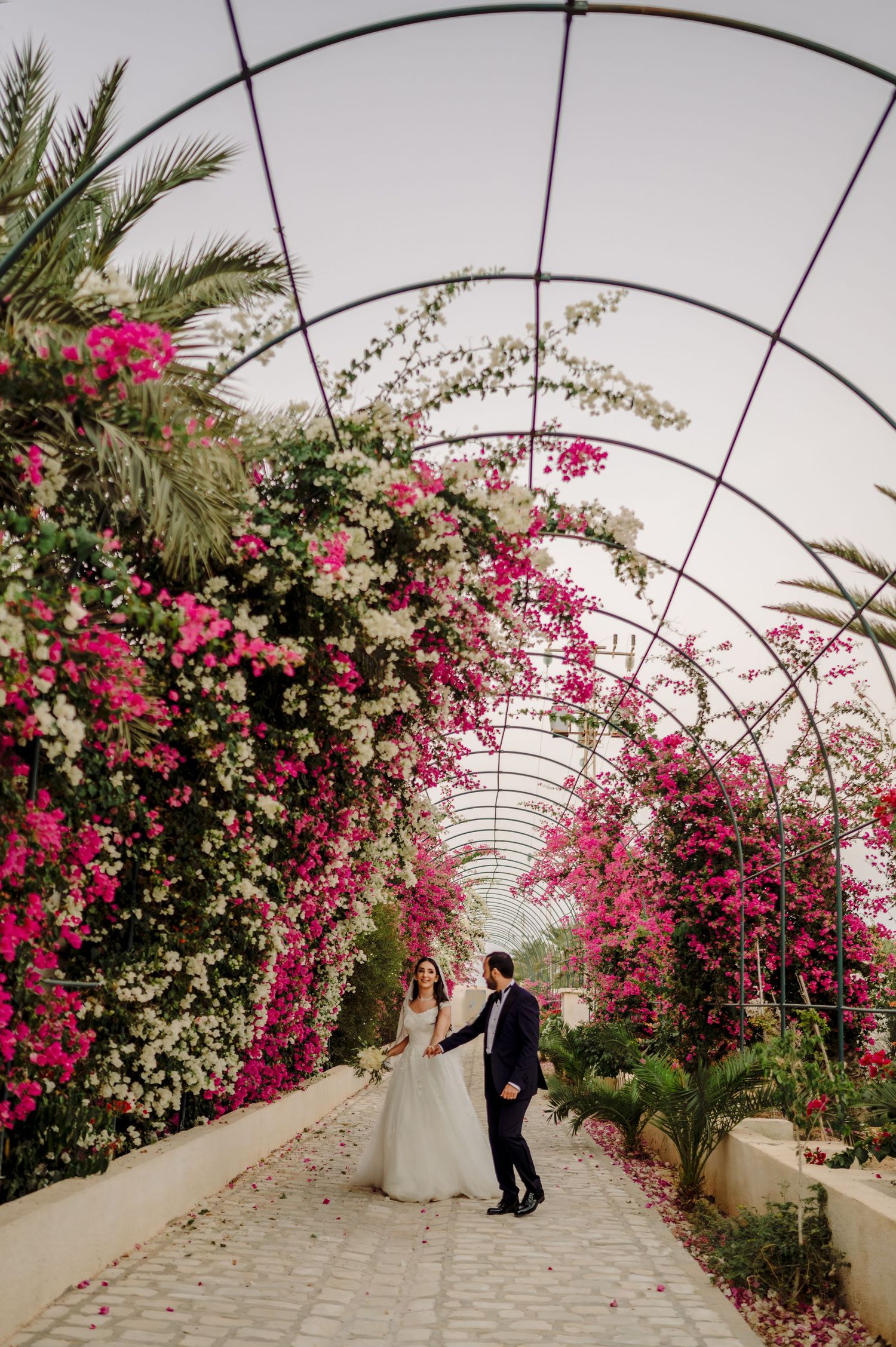 Bride and groom walking through stunning floral tunnel archway covered in pink and white bougainvillea blooms.