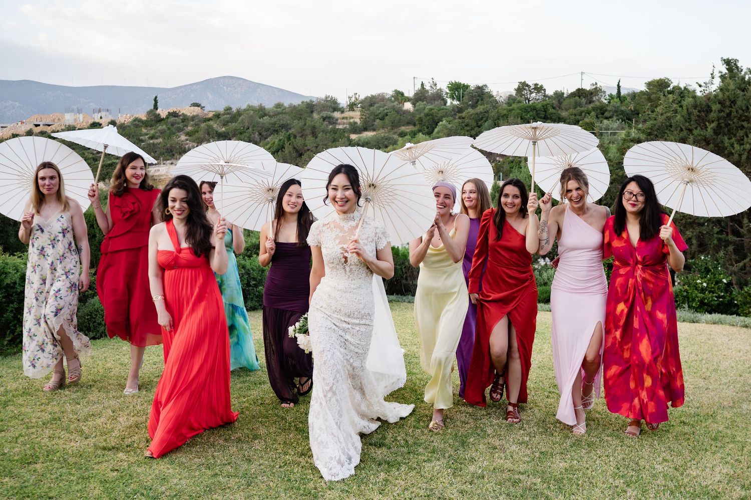Bride in lace gown poses with bridesmaids in colorful dresses holding white parasols at outdoor wedding.