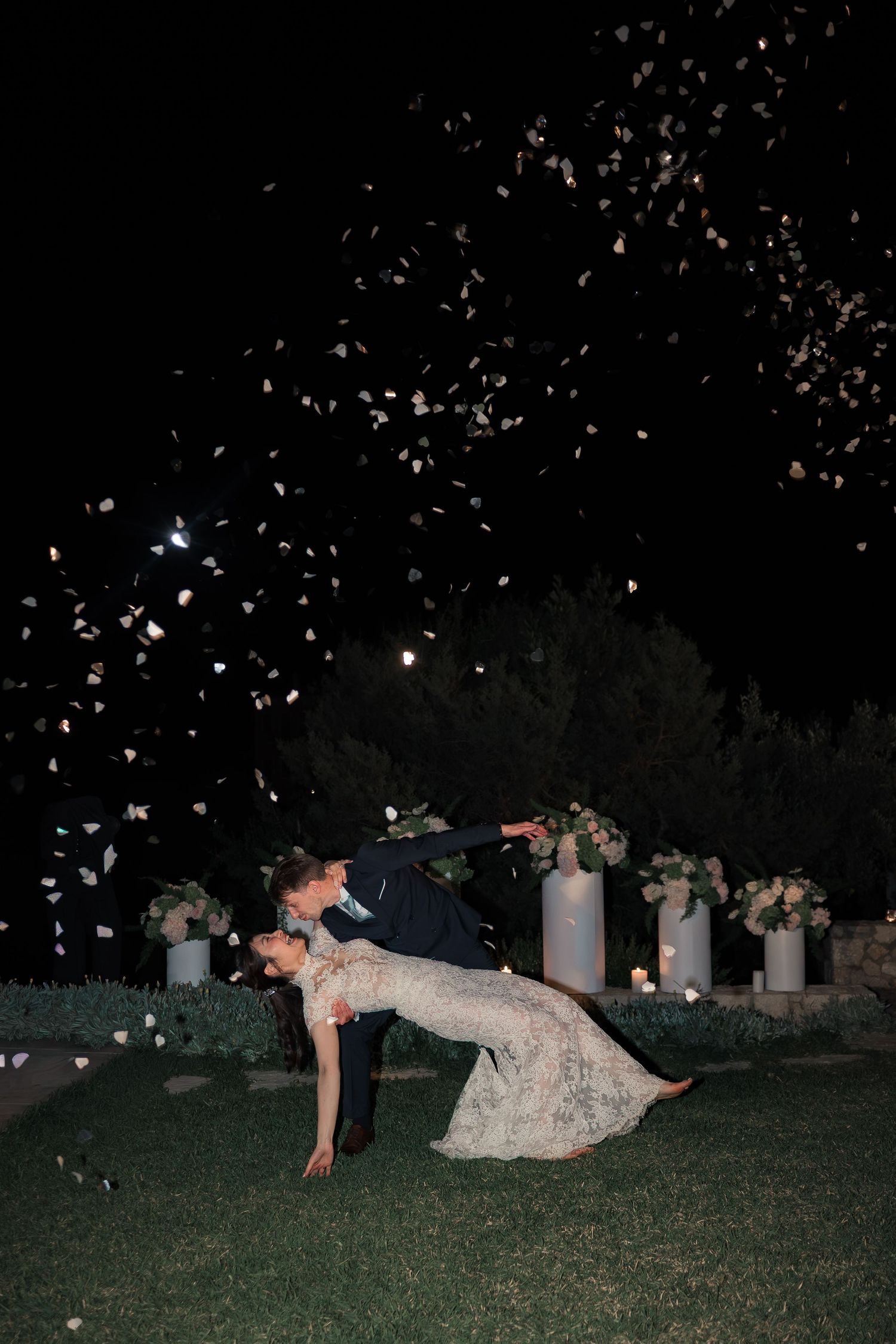 Groom dipping bride during first dance as flower petals fall at outdoor night wedding.