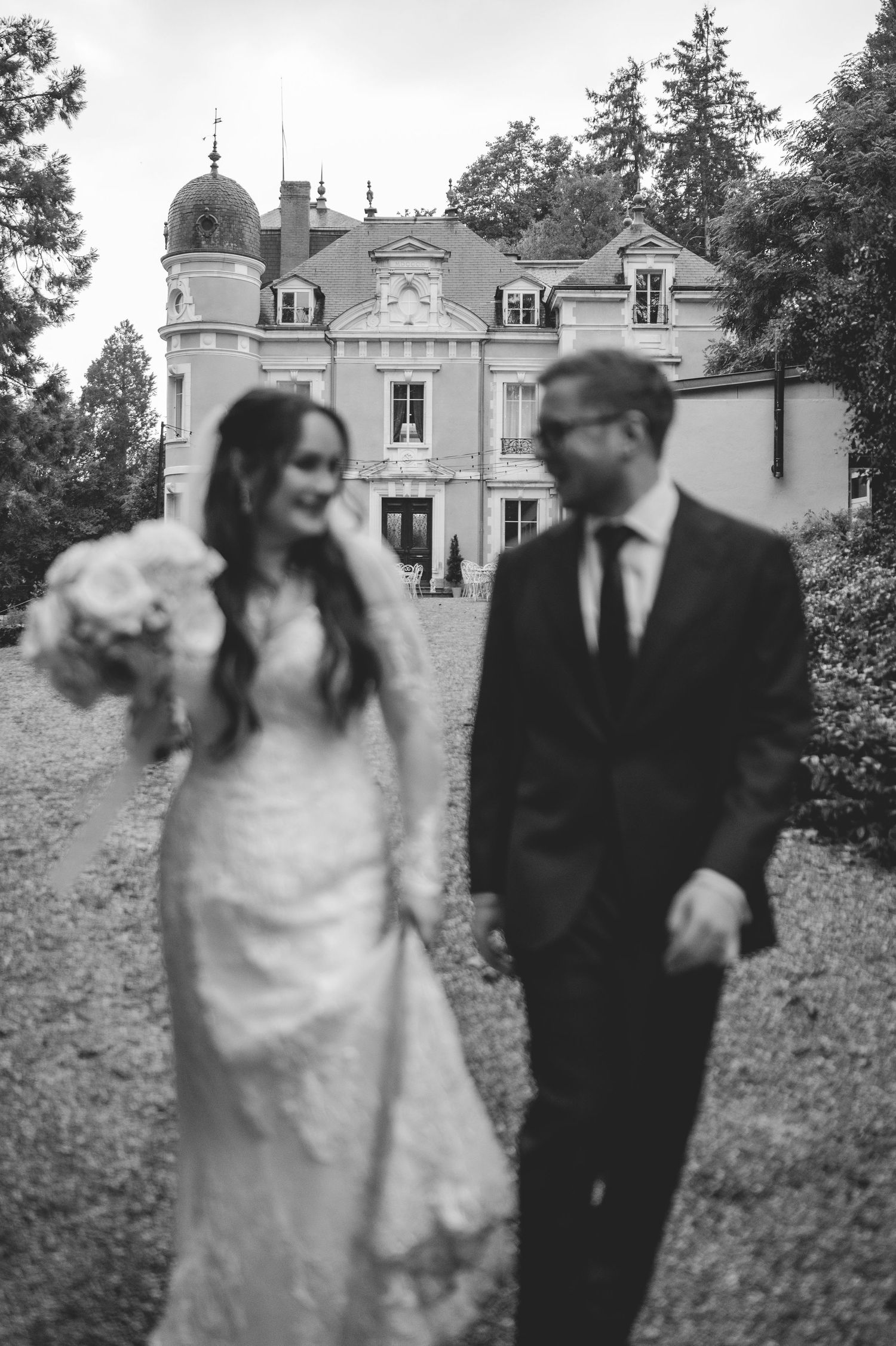 Black and white candid photo of smiling bride and groom strolling near a French chateau.