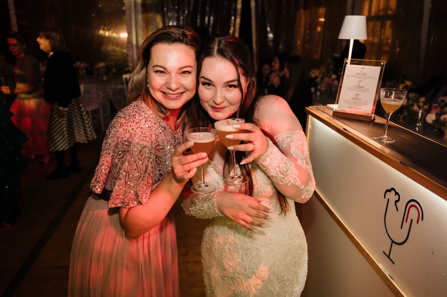 Two smiling women clink cocktail glasses together at wedding reception bar.
