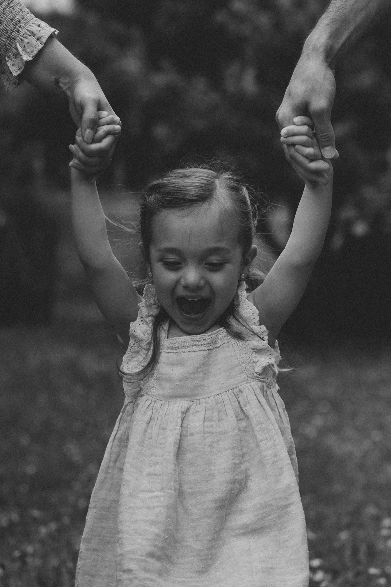 Black and white photo of a joyful little girl in a dress holding adults hands with arms raised overhead.