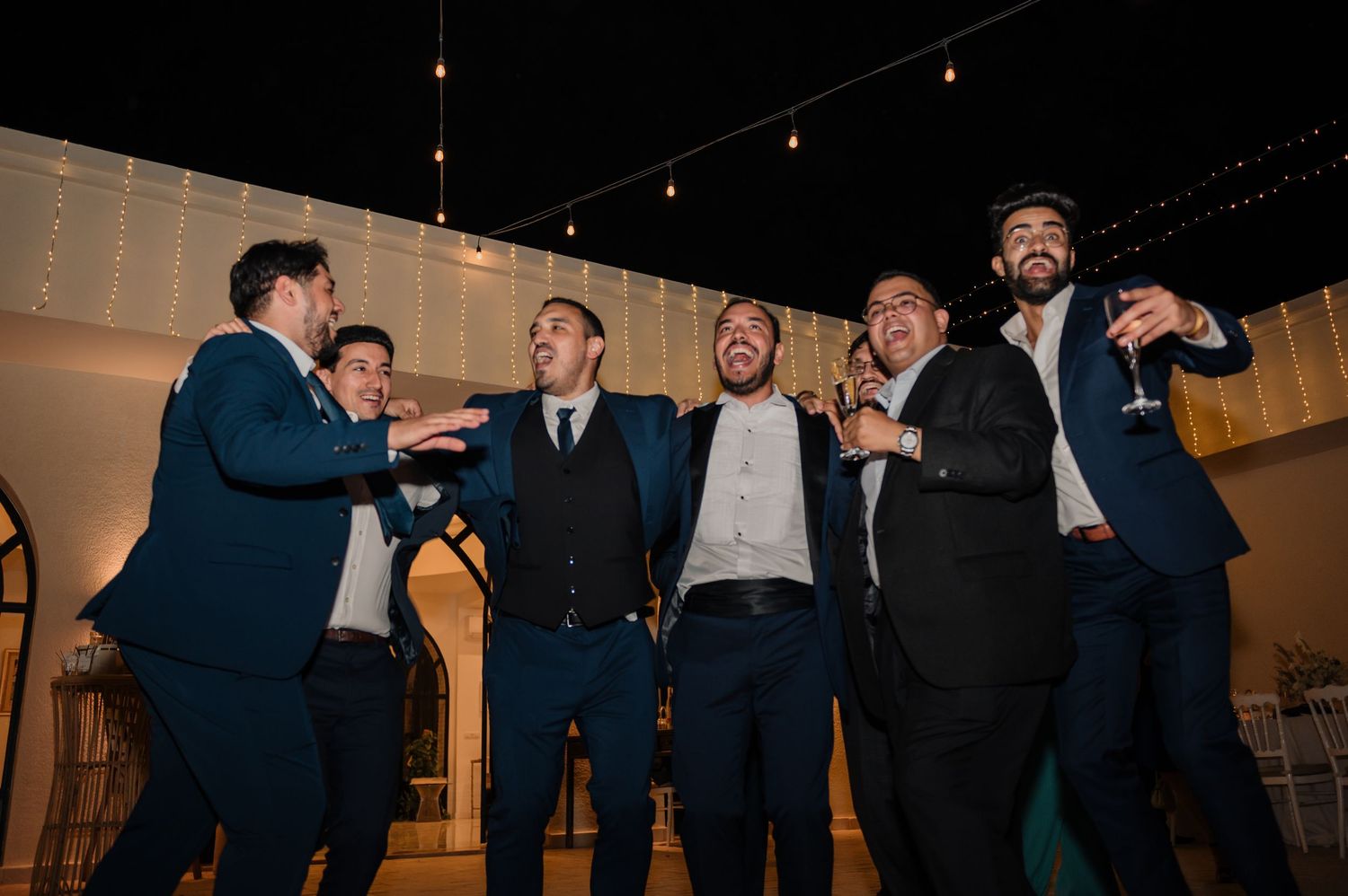 Groomsmen in navy suits laughing and celebrating together at an outdoor wedding reception at night.