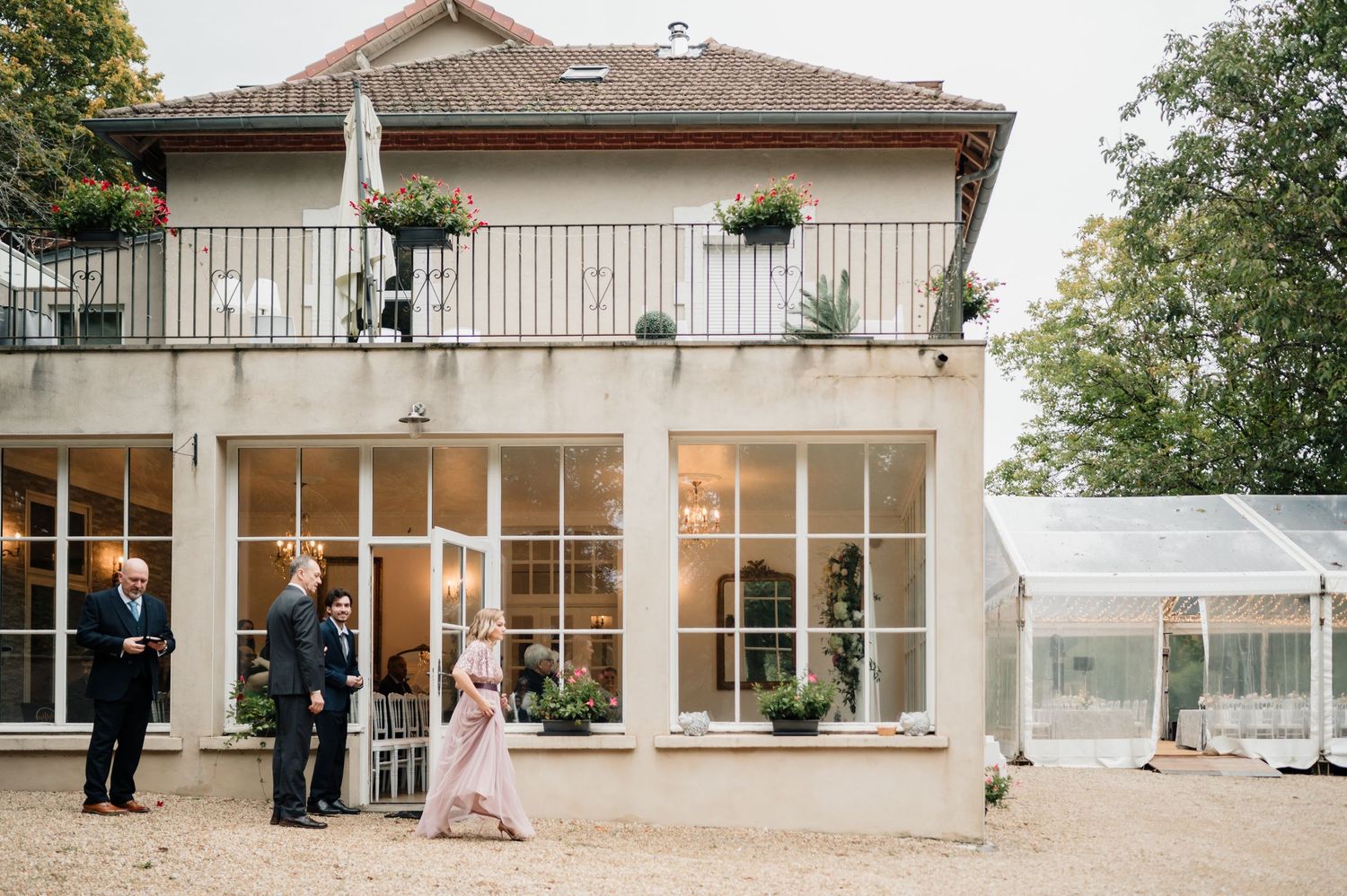 Elegant wedding venue exterior with guests mingling outside a French-style manor house with large windows and balcony.