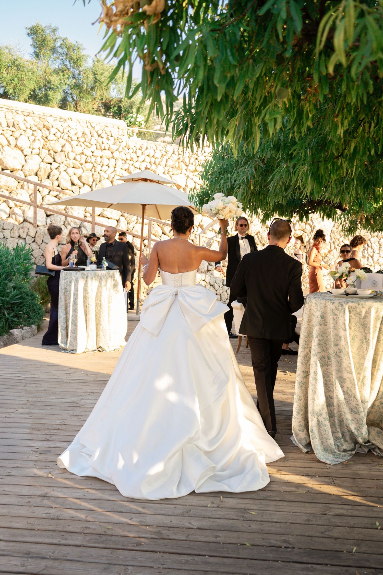 Bride in stunning white ball gown with large bow detail walking with groom at outdoor wedding reception.