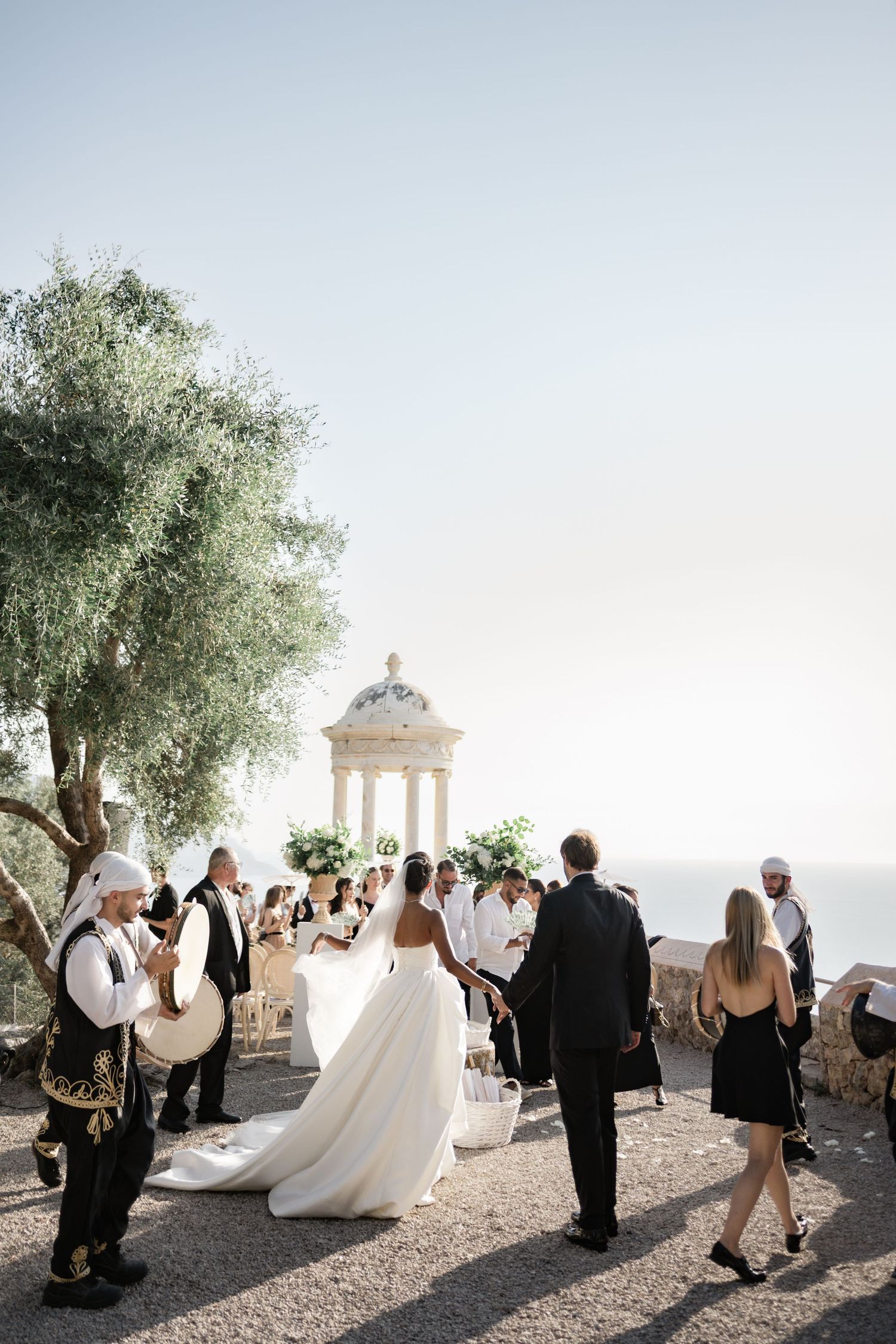 Bride walks down outdoor aisle with musicians at elegant Mediterranean wedding ceremony by the sea.