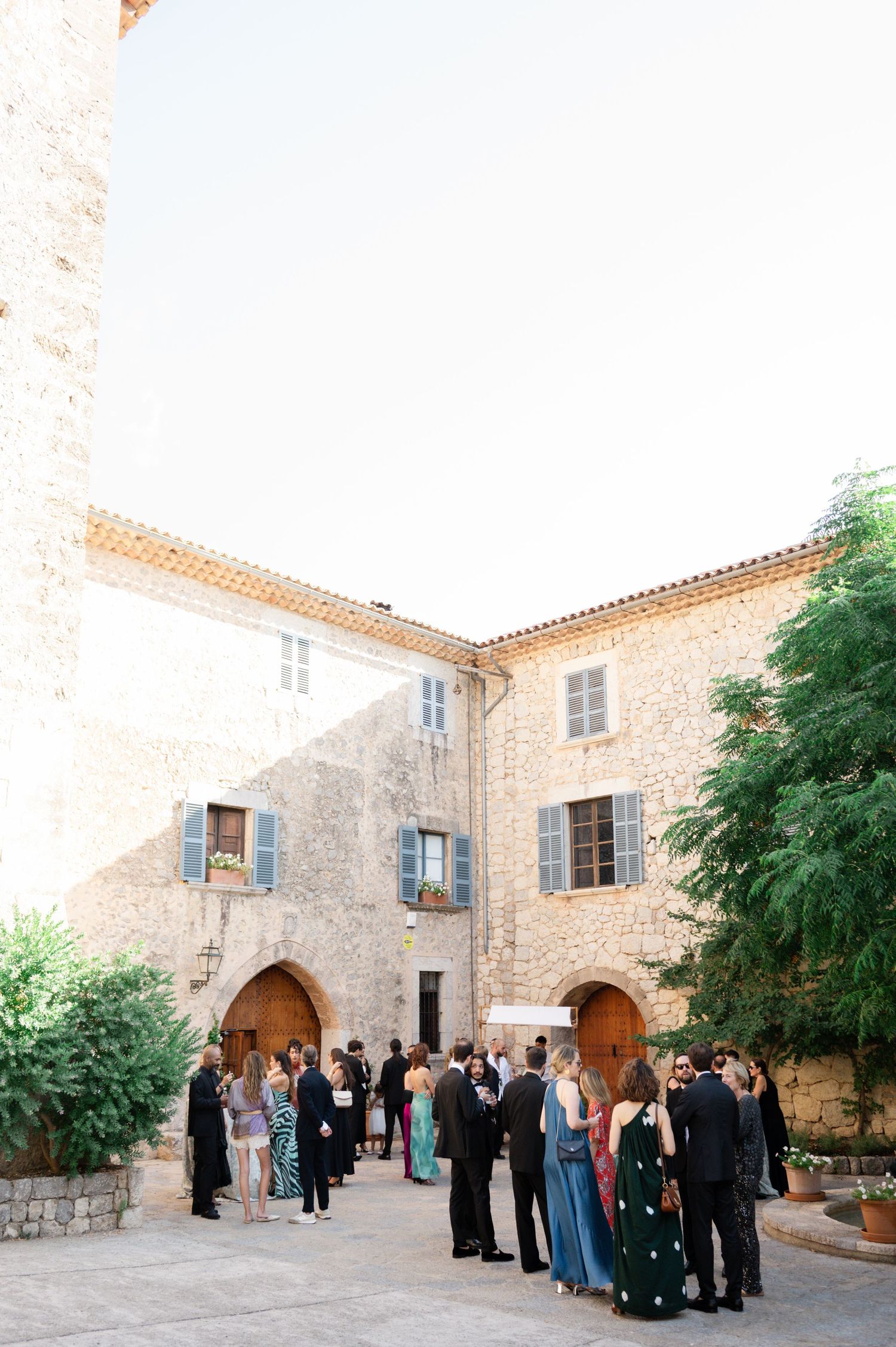Wedding guests in formal evening wear gather in a courtyard of a historic stone venue at dusk.