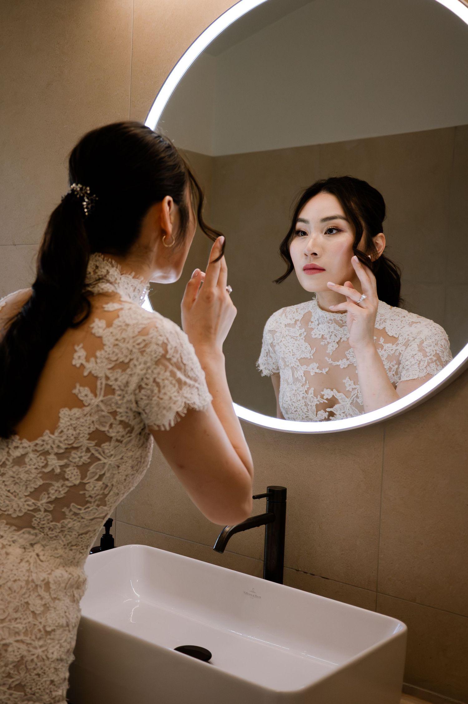 Bride in lace wedding dress checking makeup in a round illuminated bathroom mirror before her wedding ceremony.