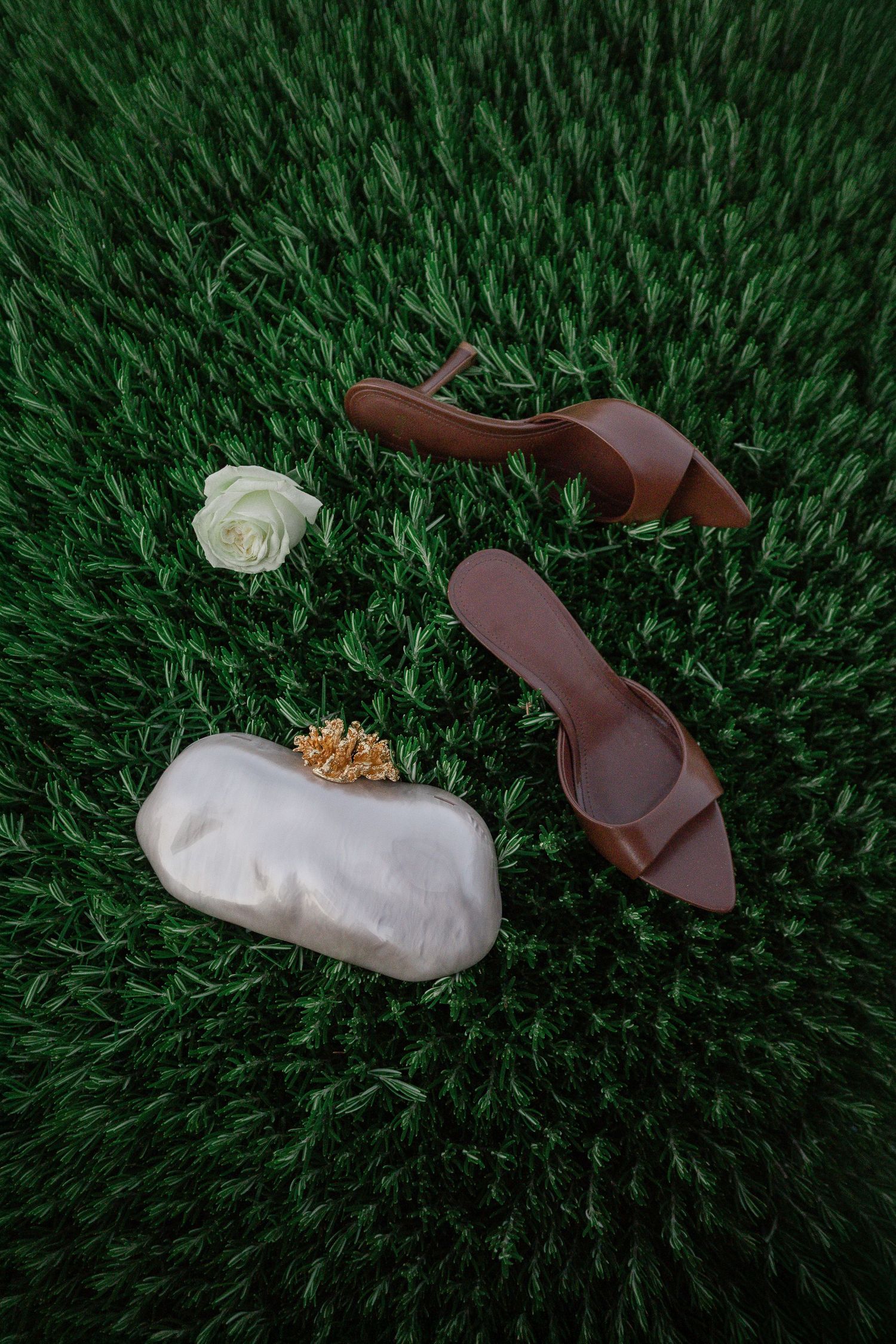 Flat lay of brown strappy heels, white pearl clutch, and a white rose on lush green grass.