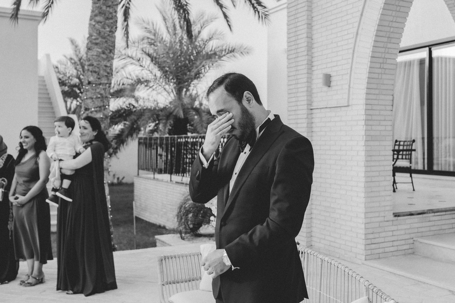 Emotional groom in black tuxedo covering his mouth as he sees his bride approaching at an outdoor wedding.