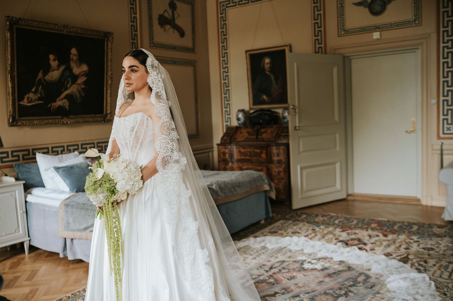 Elegant bride in long-sleeve lace wedding gown and veil holding bouquet in classical portrait-lined room.