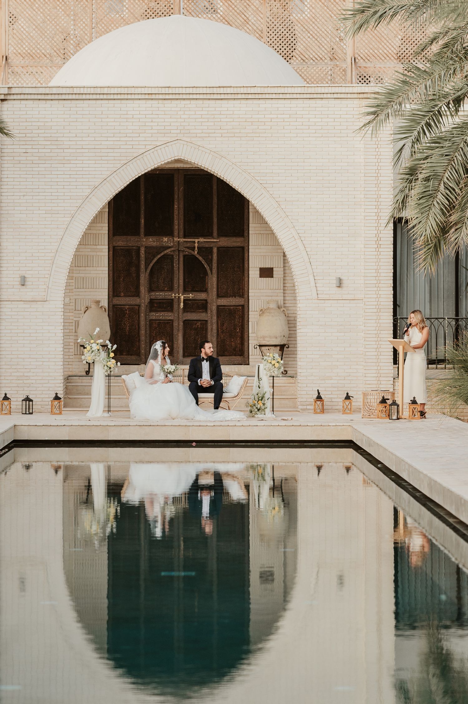 Bride and groom seated at altar reflected in pool before grand arched doorway at luxury wedding venue.