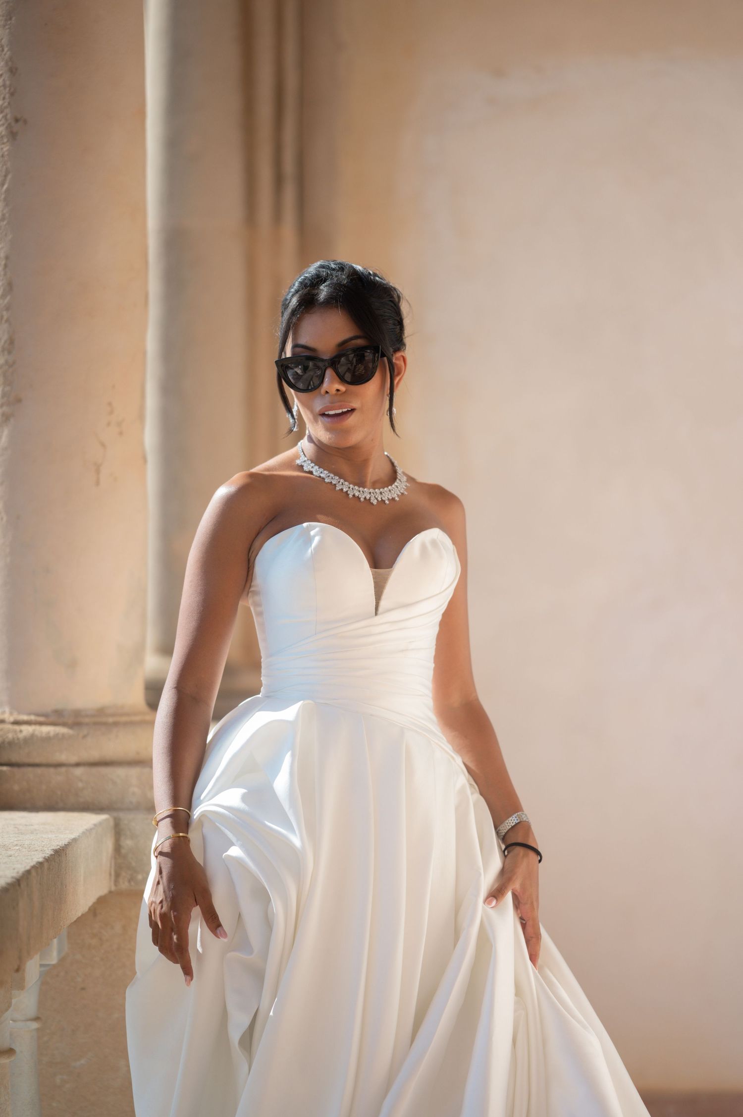 Stylish bride in strapless white gown and sunglasses with diamond necklace poses by stone columns.