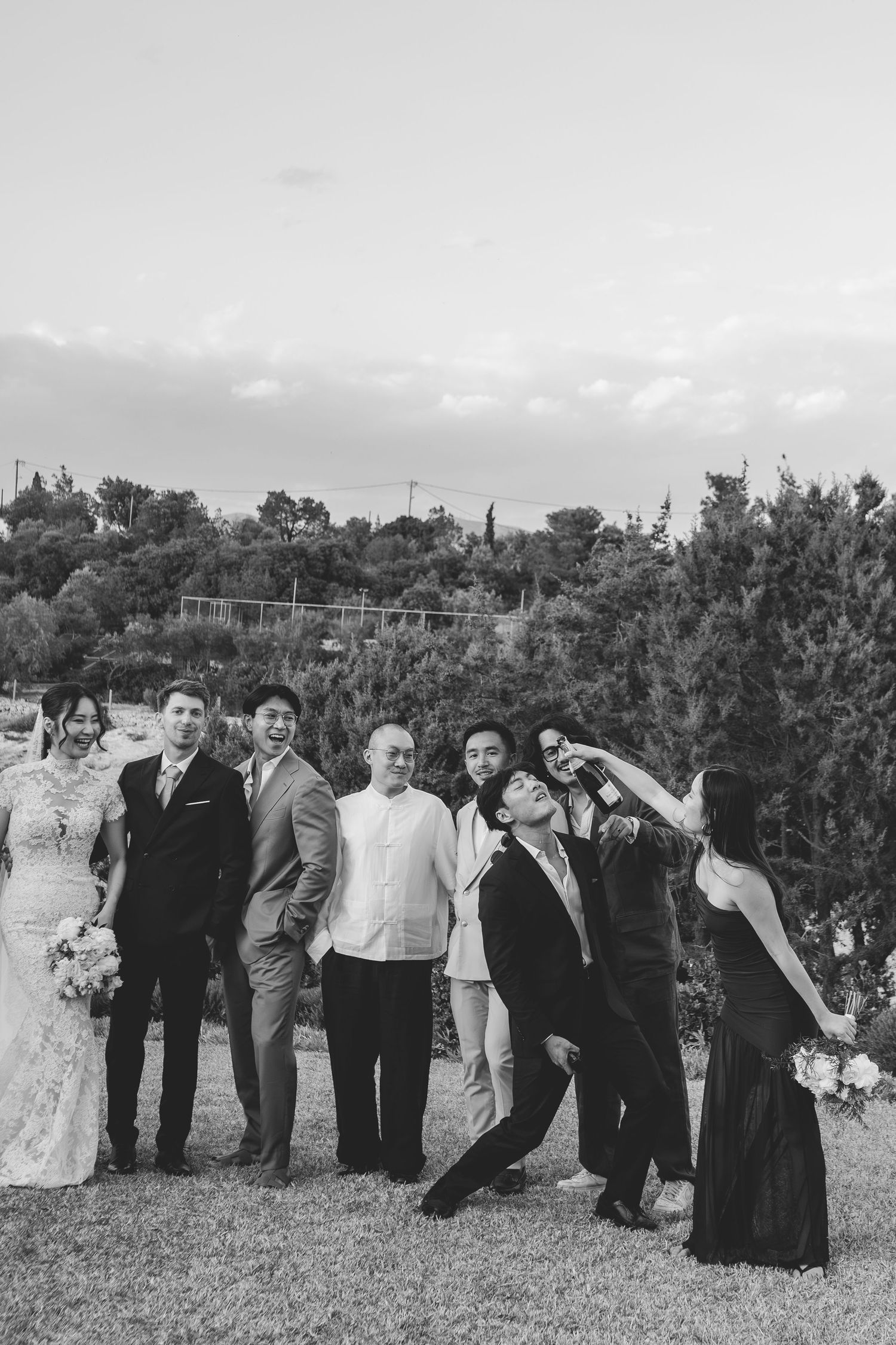Wedding party posing casually on lawn with trees in black and white photography.