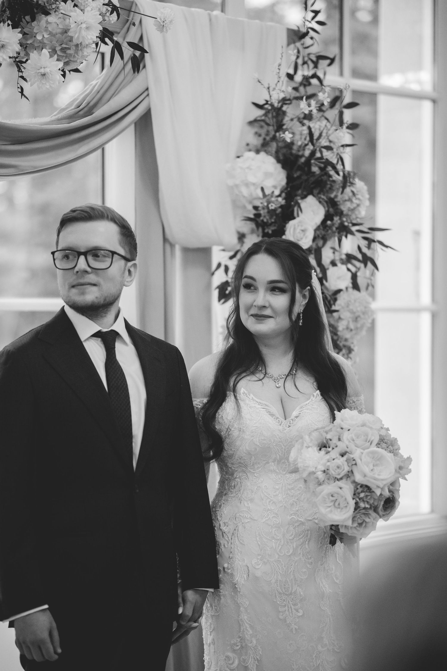 Black and white portrait of a bride and groom standing at the altar with floral arrangements.