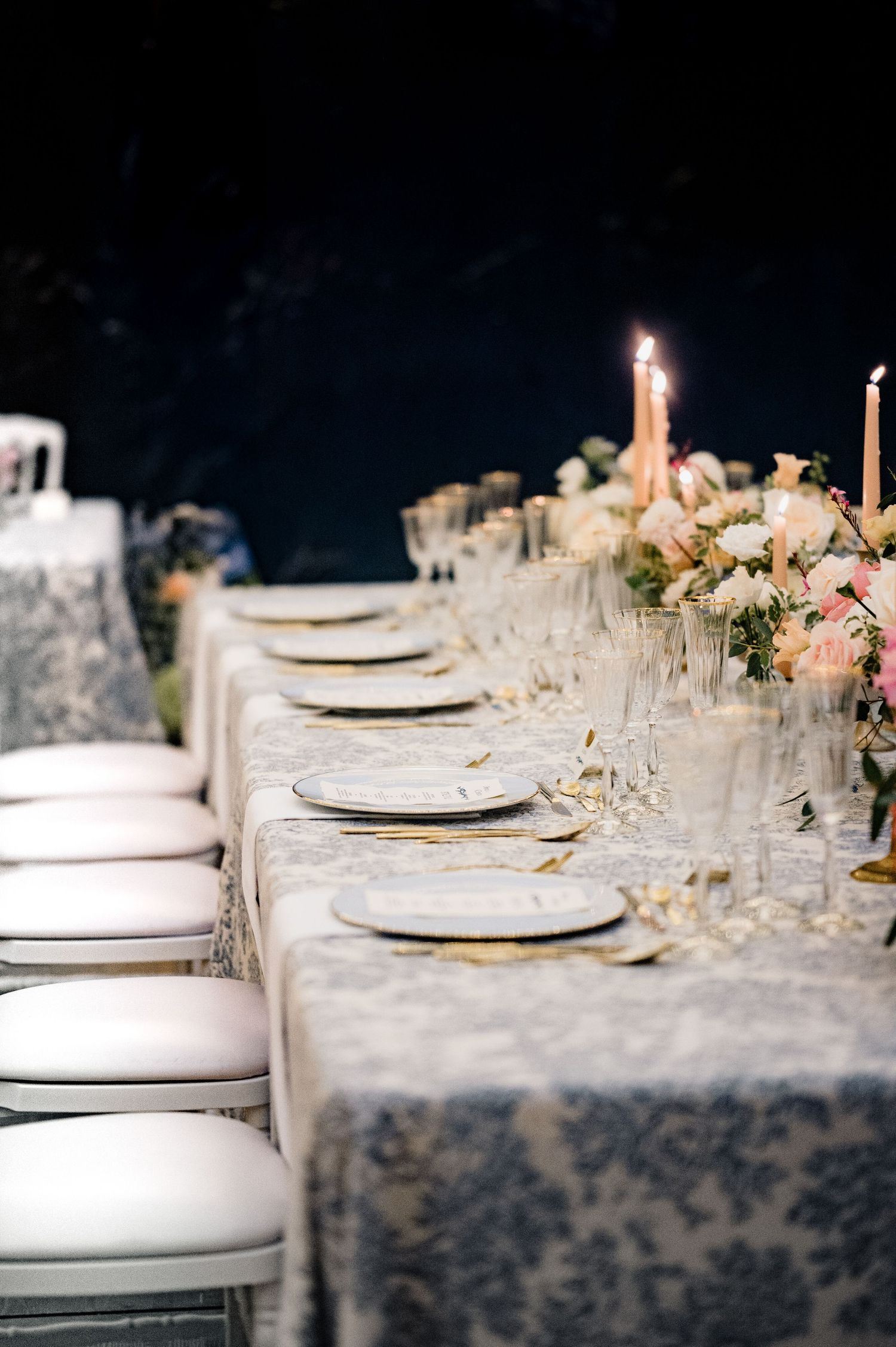 Long elegant wedding banquet table with blue toile linen, white plates, gold cutlery, and glowing taper candles.