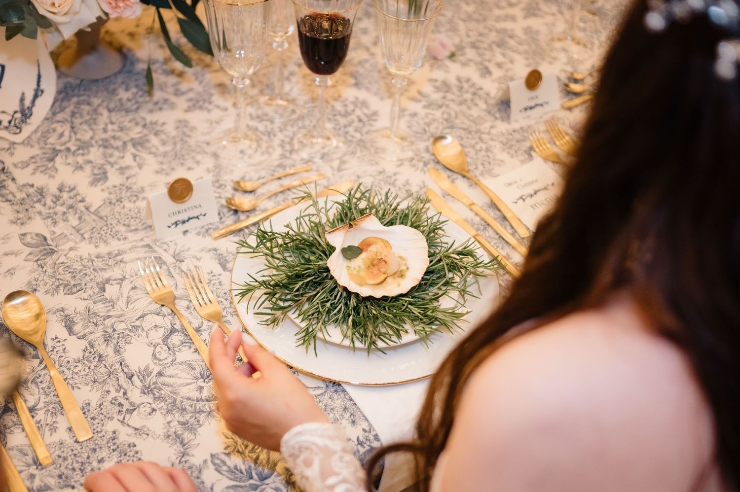 Bride seated at a blue toile wedding table with a scallop shell dish and gold cutlery.