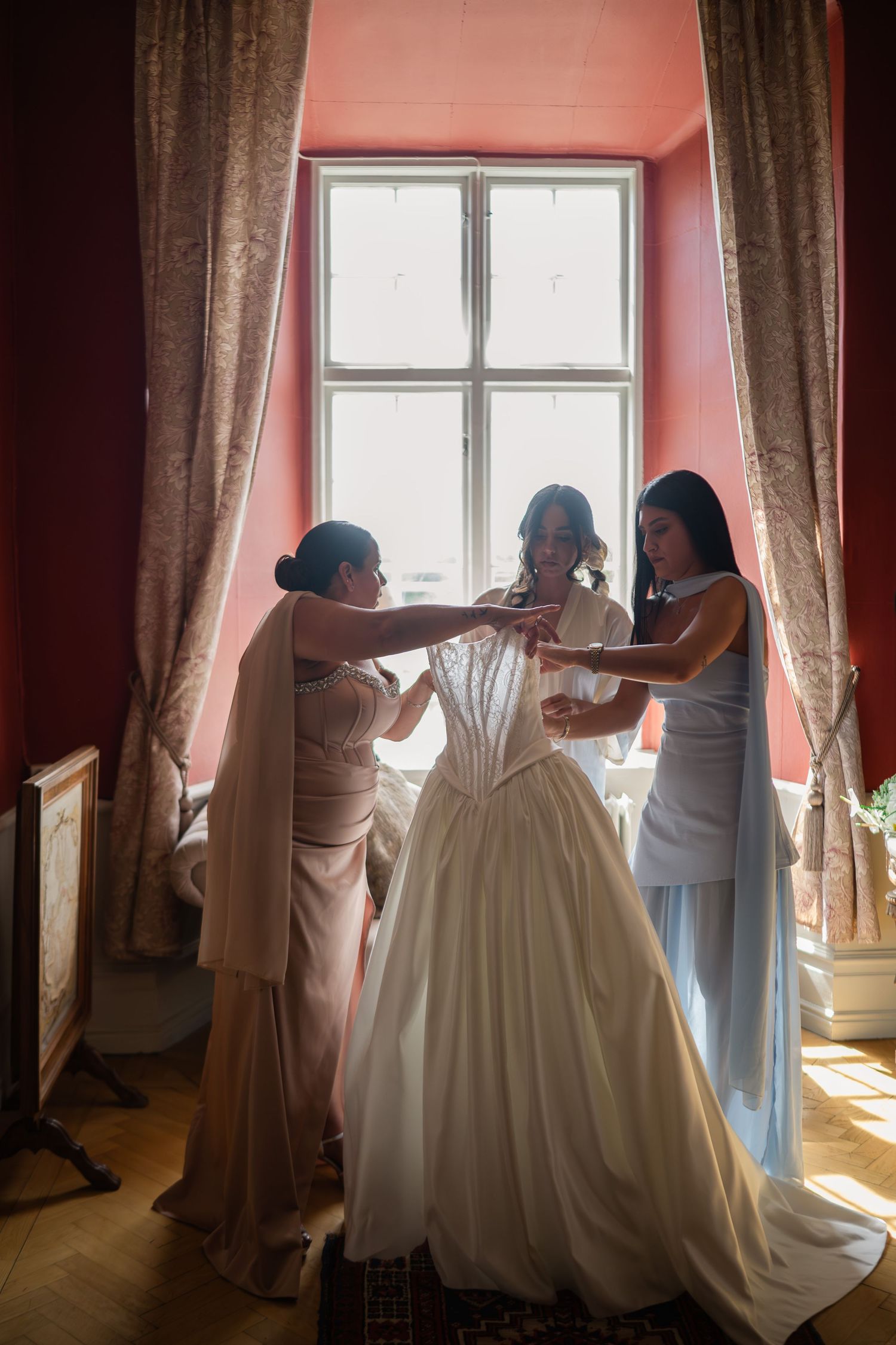 Two bridesmaids in blue gowns helping bride into lace corset wedding dress by pink-walled window.