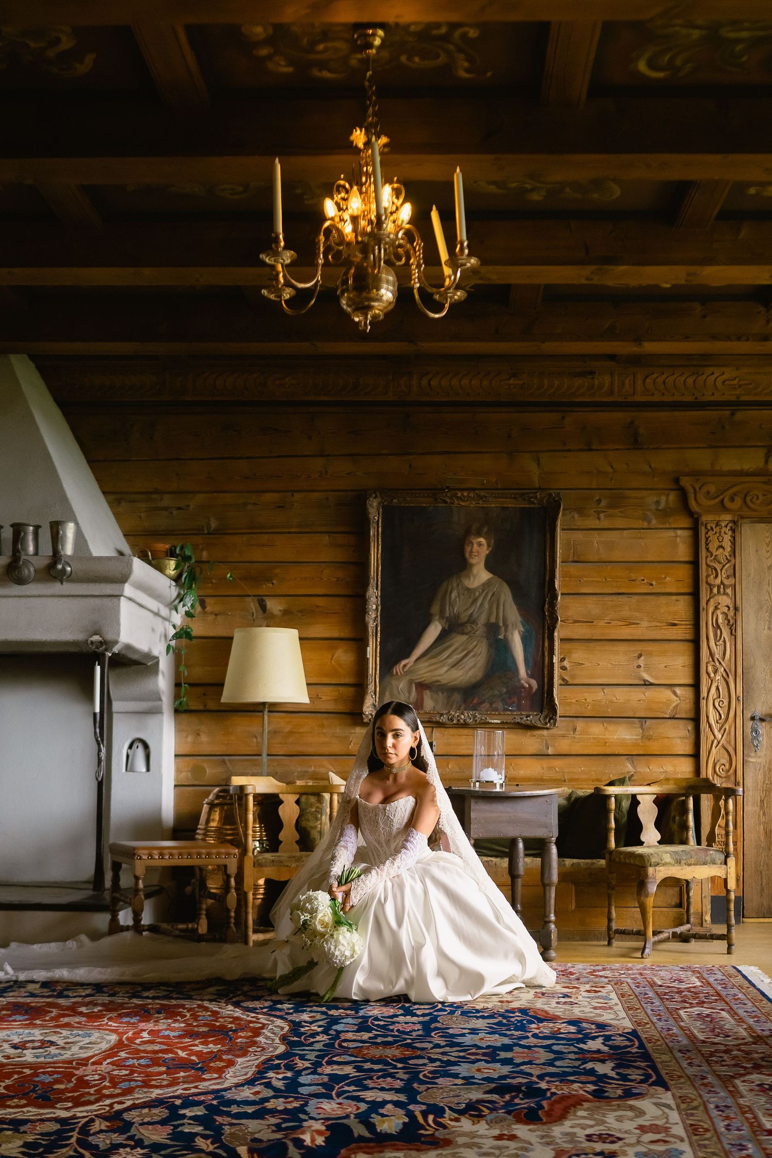 Bride in white ball gown seated in rustic wooden room with chandelier and vintage portrait.