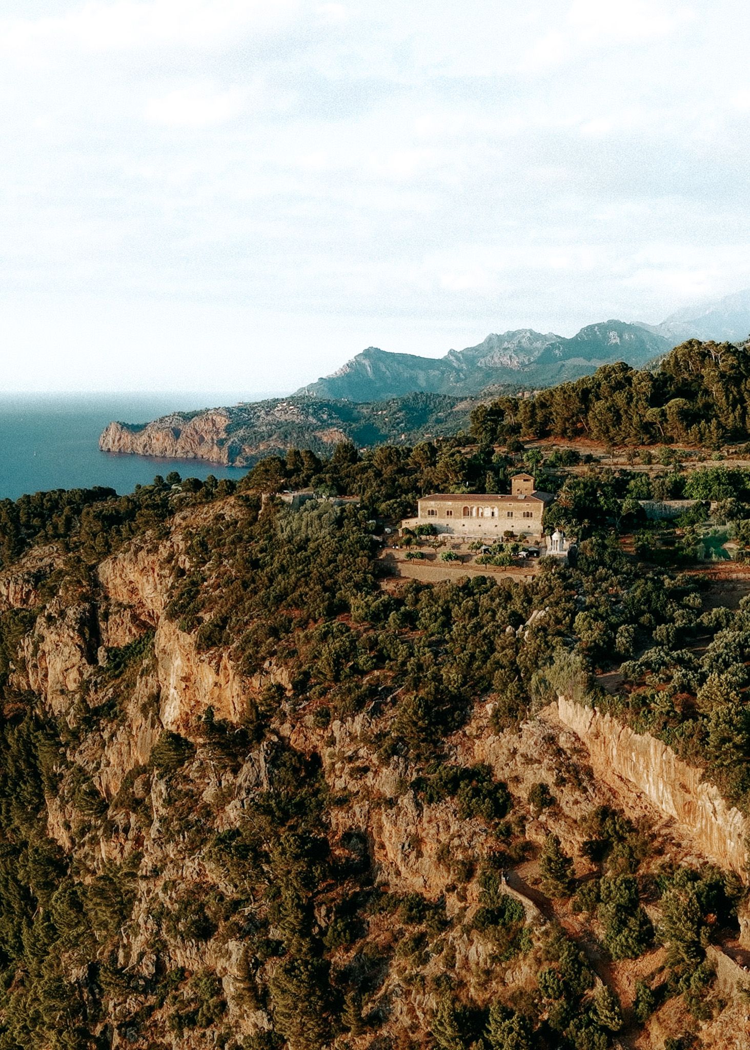 A secluded stone farmhouse sits on a rocky cliff surrounded by Mediterranean greenery with sea views.