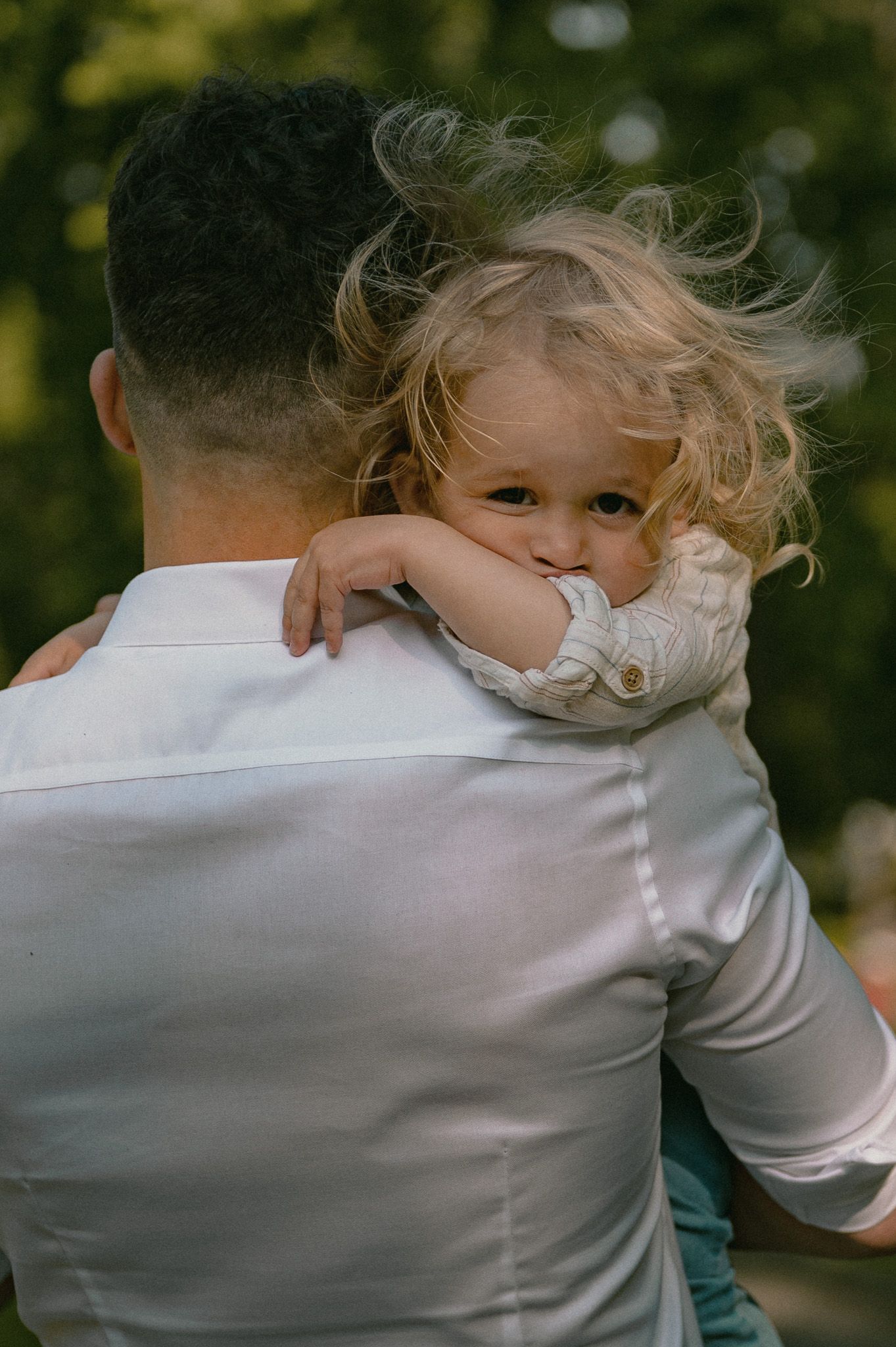 Tender moment of a curly-haired toddler resting head on father's shoulder while being carried outdoors.