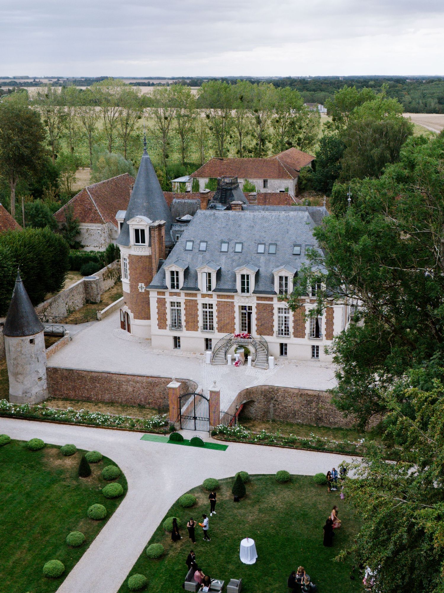Aerial view of a stunning French château with turrets, brick facade, and manicured gardens surrounded by lush countryside.