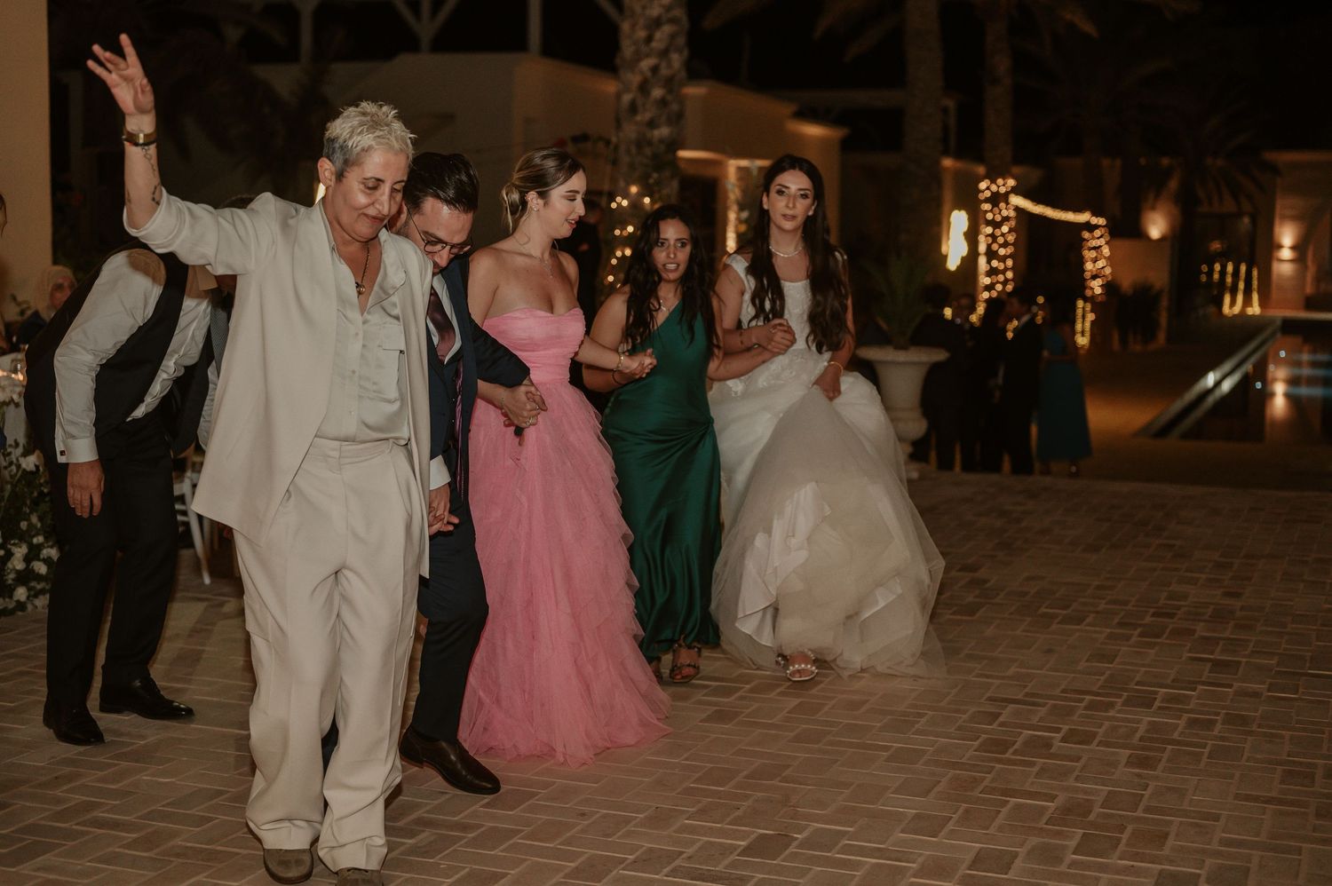 Wedding guests dancing outdoors at night with string lights, bride in white gown and guests in colorful attire.