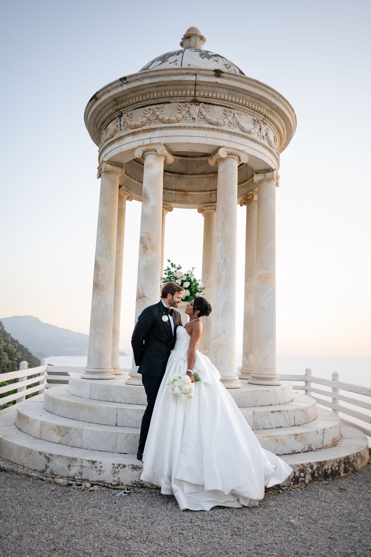 Bride and groom share romantic kiss under ornate marble rotunda with sea views at sunset wedding.