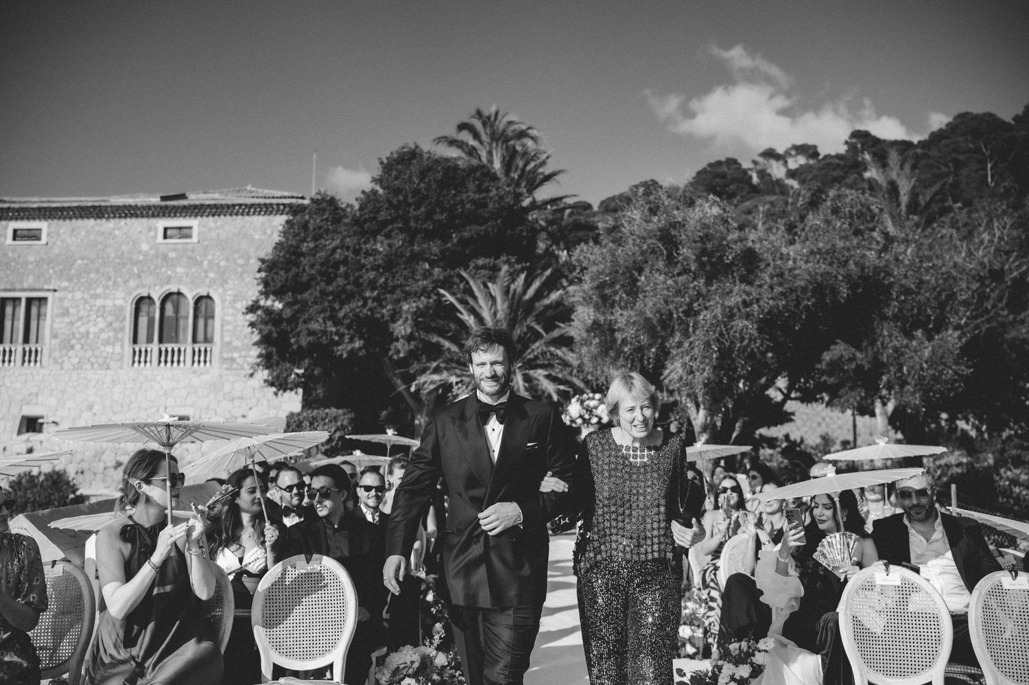 Black and white photo of a groom walking down the aisle with a guest at an outdoor wedding.