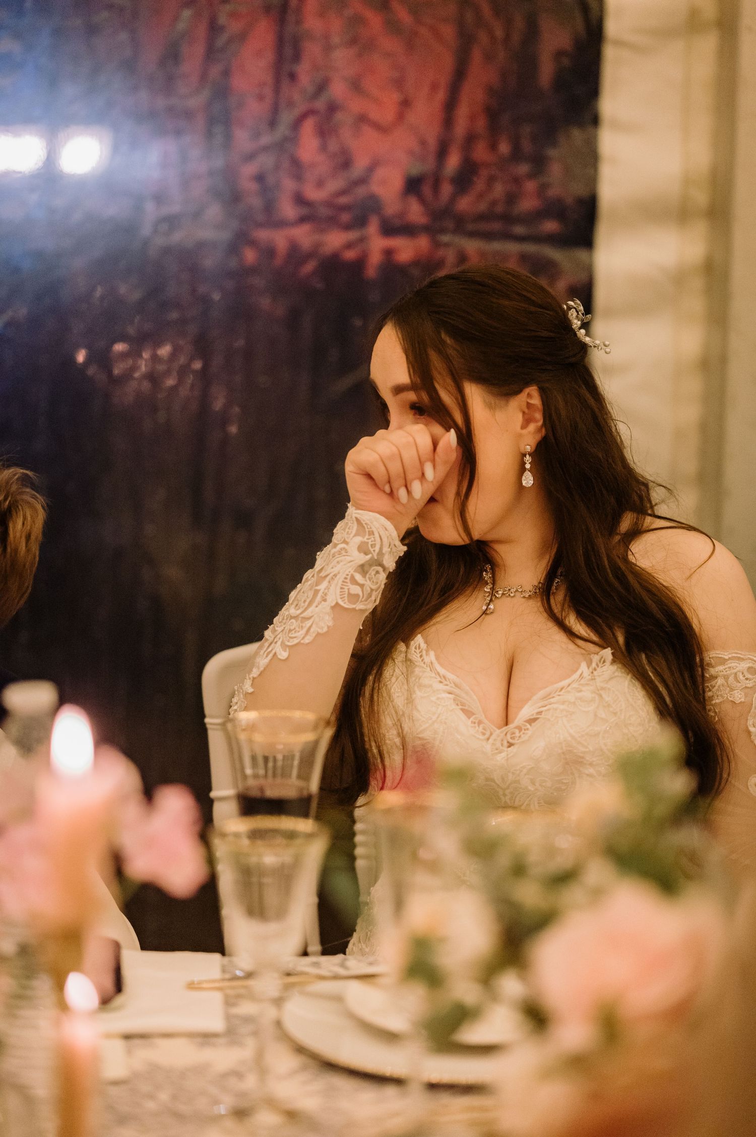 Emotional bride in lace wedding dress covering her mouth during heartfelt wedding reception speeches.