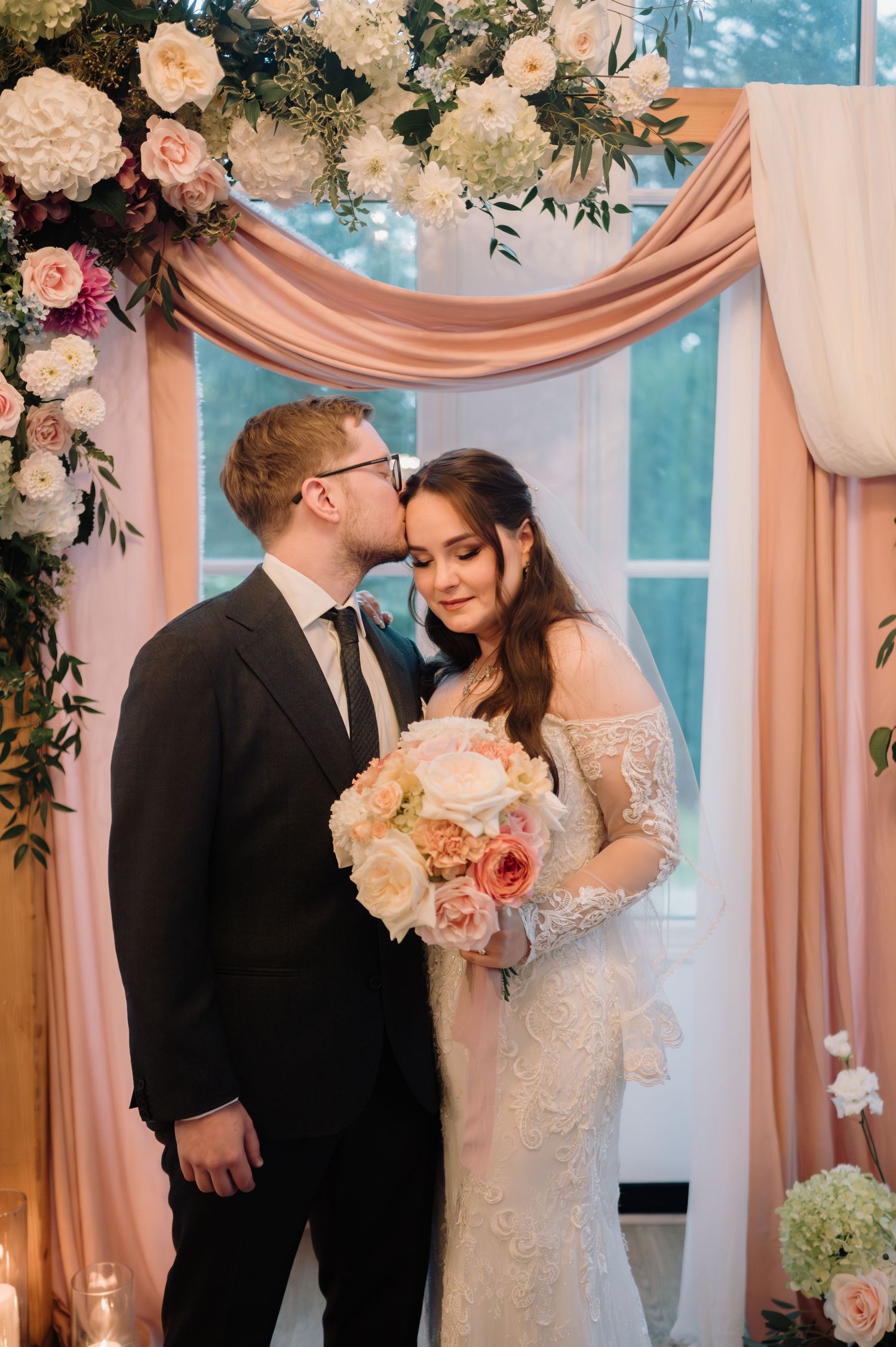Groom kisses bride on the cheek as she smiles holding a peach rose bouquet at the altar.