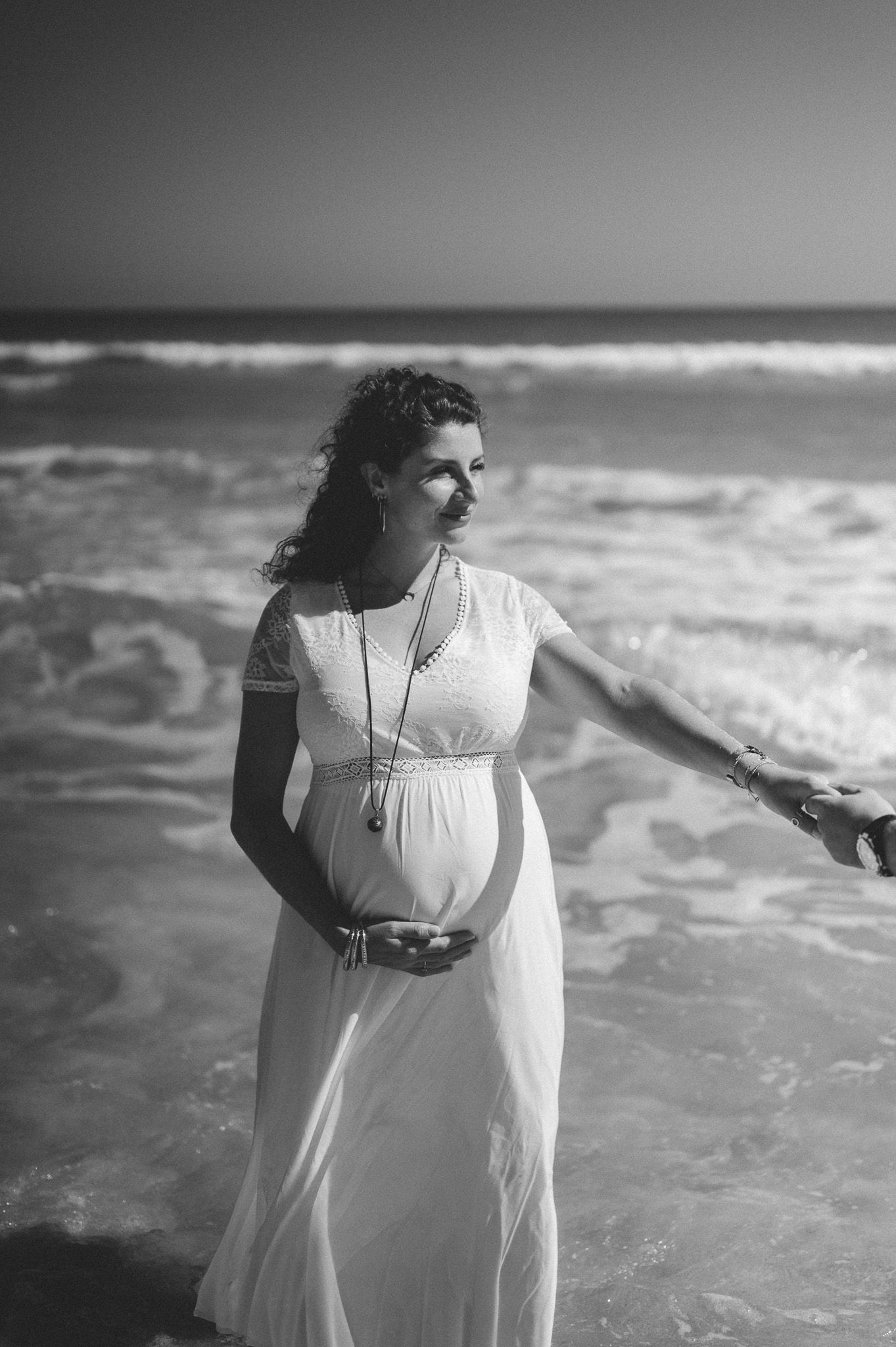 Pregnant woman in white dress cradles belly on beach, black and white maternity photo with ocean waves.