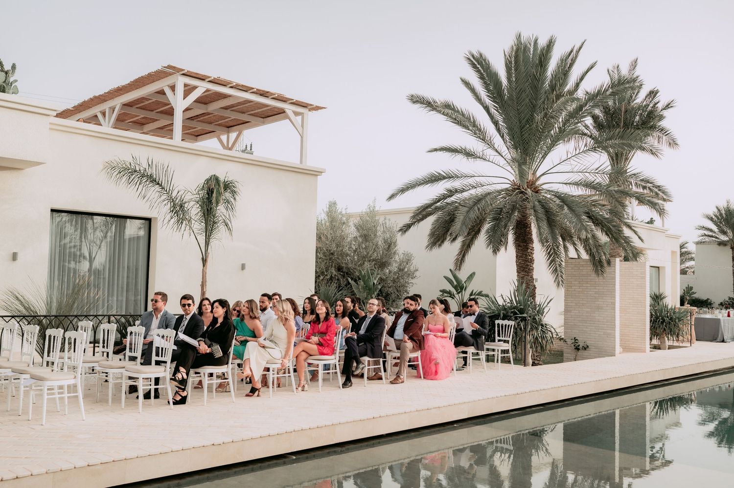 Wedding guests seated in white chairs beside pool with palm trees at Moroccan villa.
