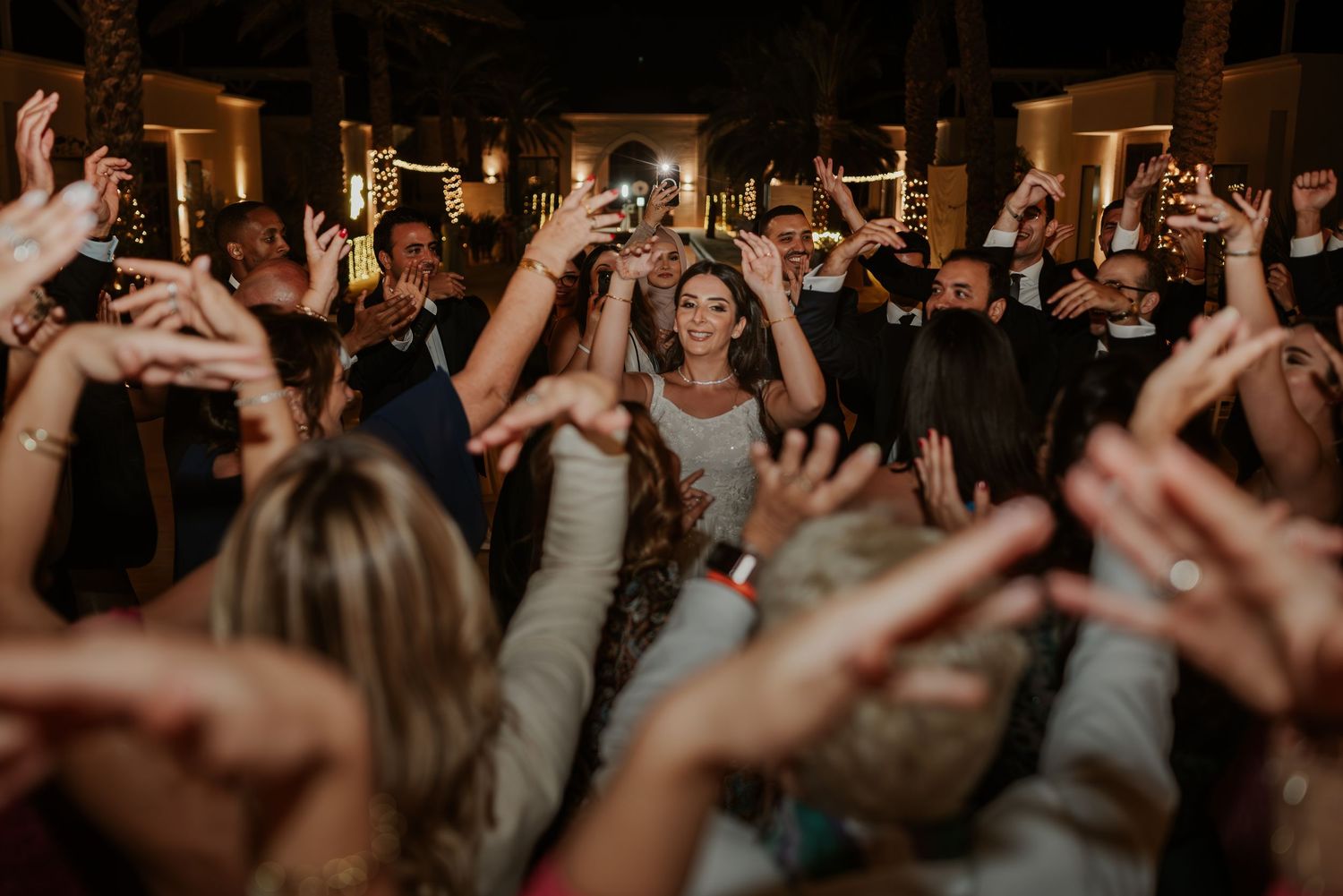 Bride walks through crowd of cheering guests with raised hands at outdoor wedding reception.