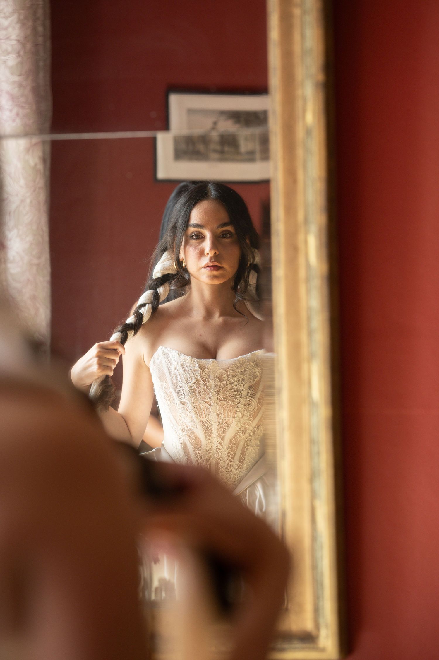 Bride in white lace corset braiding her dark hair while gazing at her reflection in an ornate gold mirror.