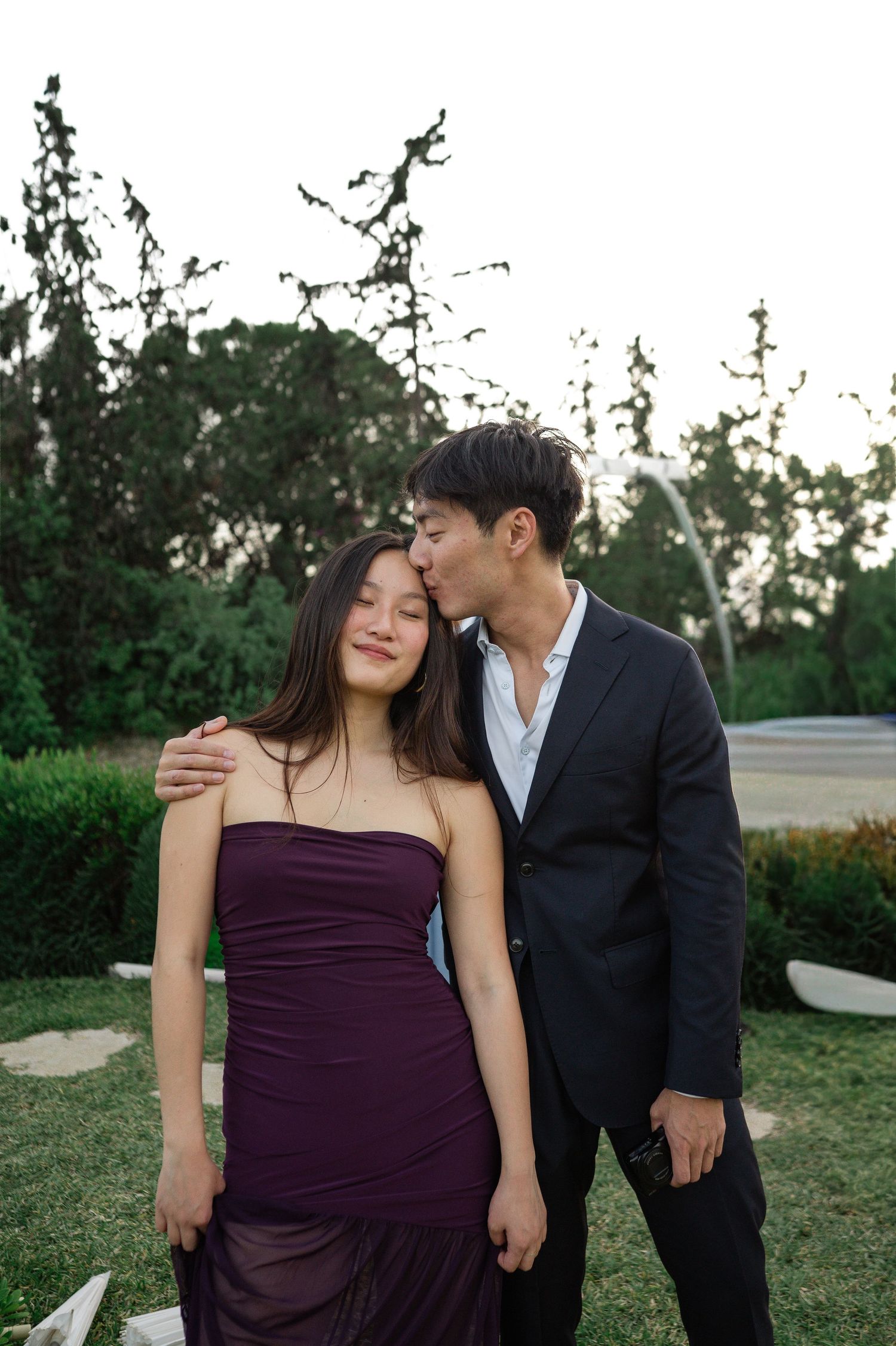 Young Asian couple sharing tender moment outdoors at wedding, man in suit kissing woman on forehead.