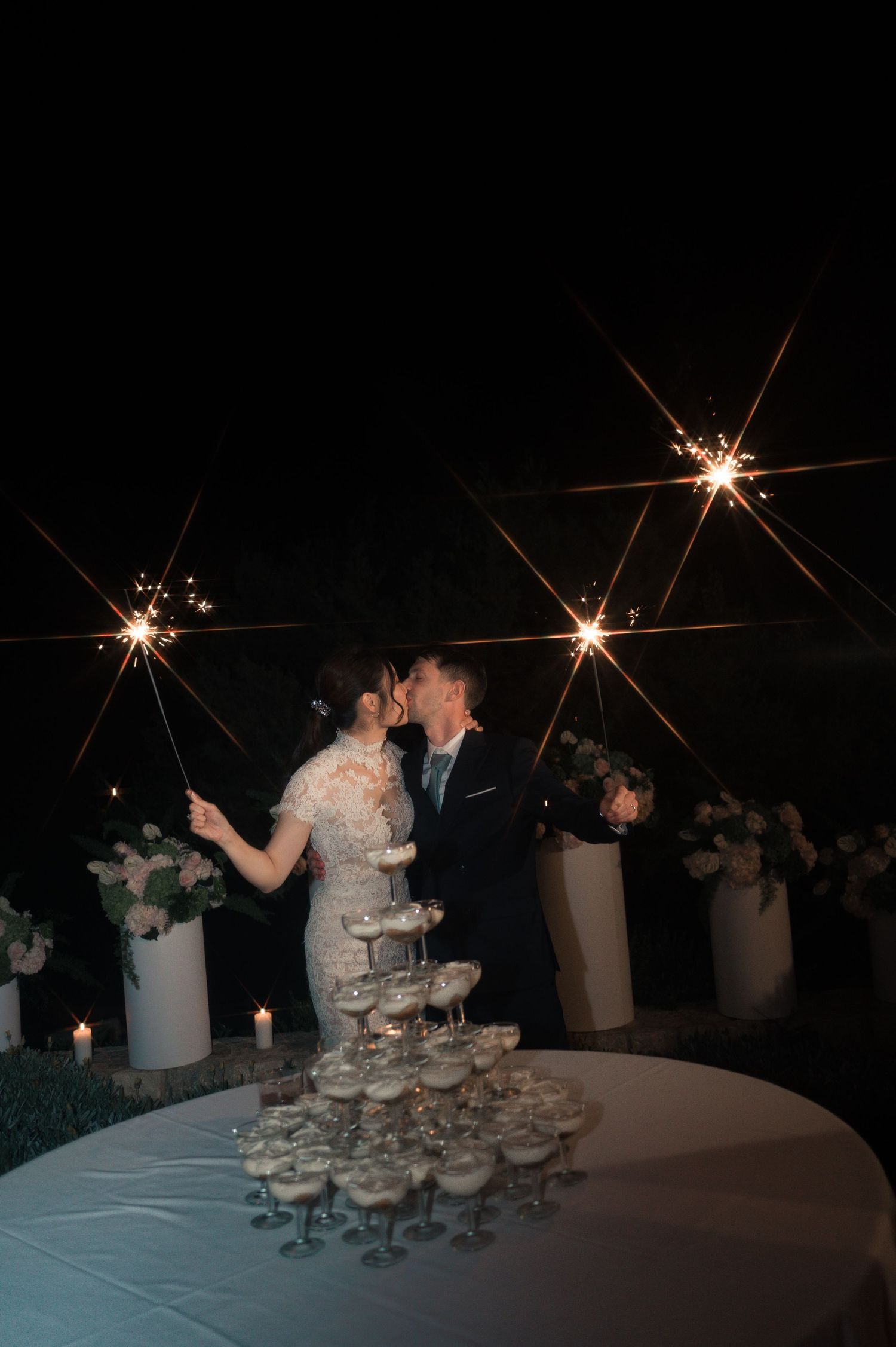 Bride and groom kissing behind a champagne tower while holding sparklers at their wedding reception.
