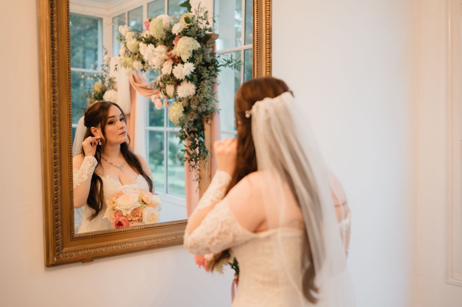 Bride in lace wedding gown and veil adjusts earring while looking in an ornate mirror.