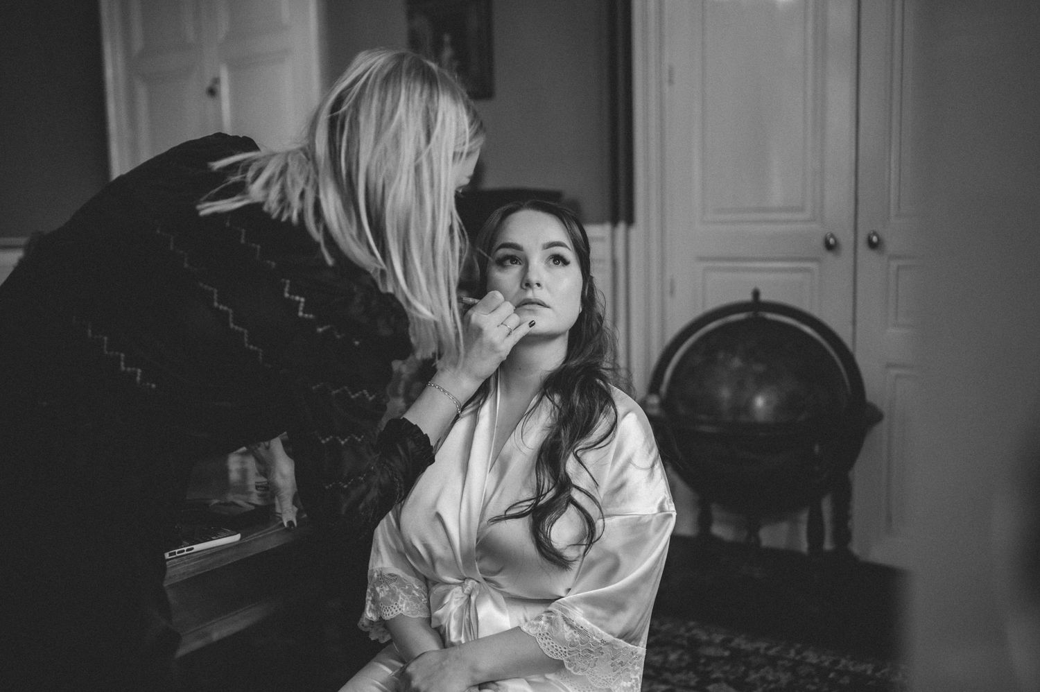 Black and white photo of a makeup artist applying bridal makeup to a bride in a satin robe.