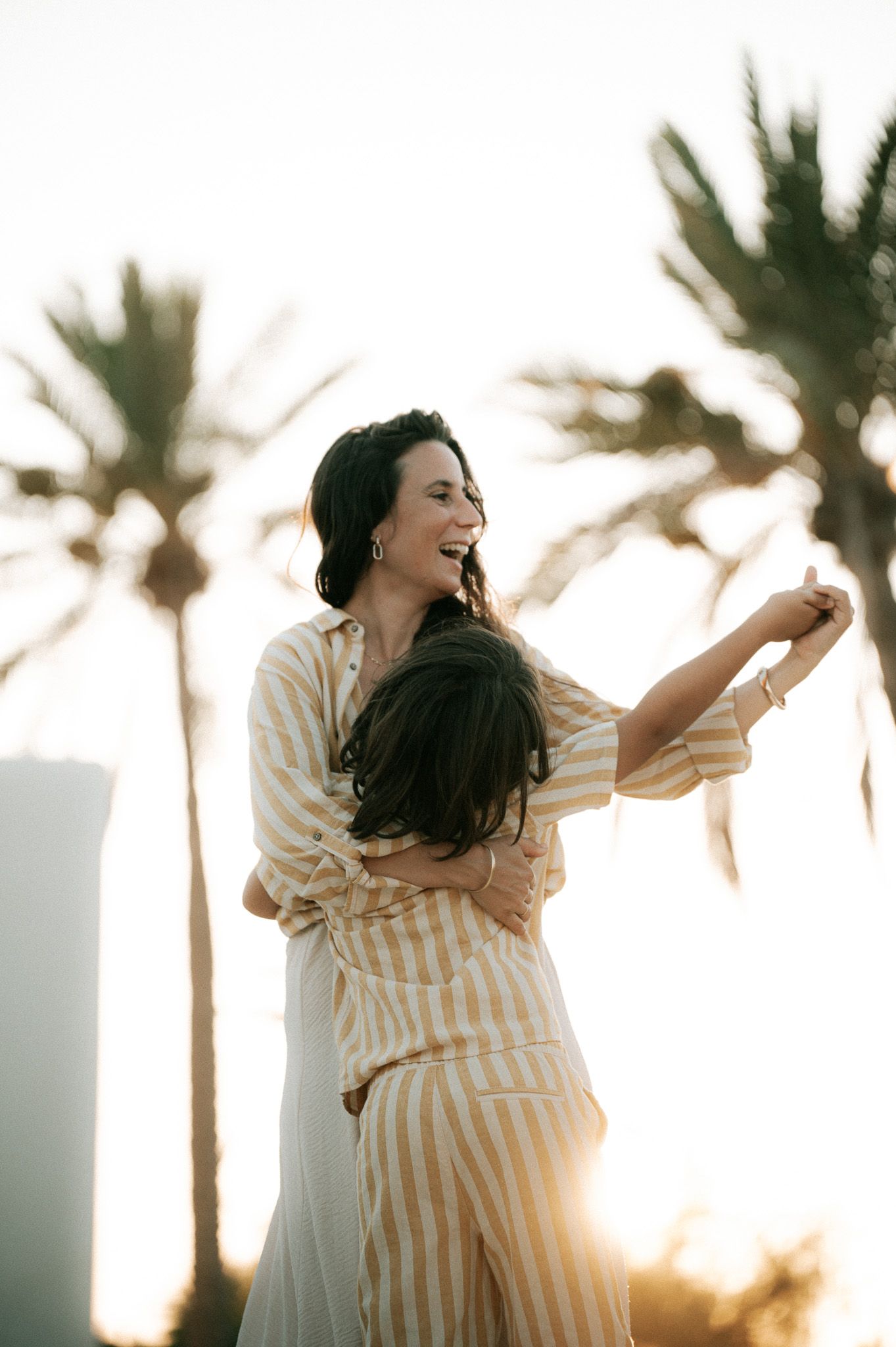 Laughing mother in striped outfit spinning and hugging her son near palm trees at sunset.