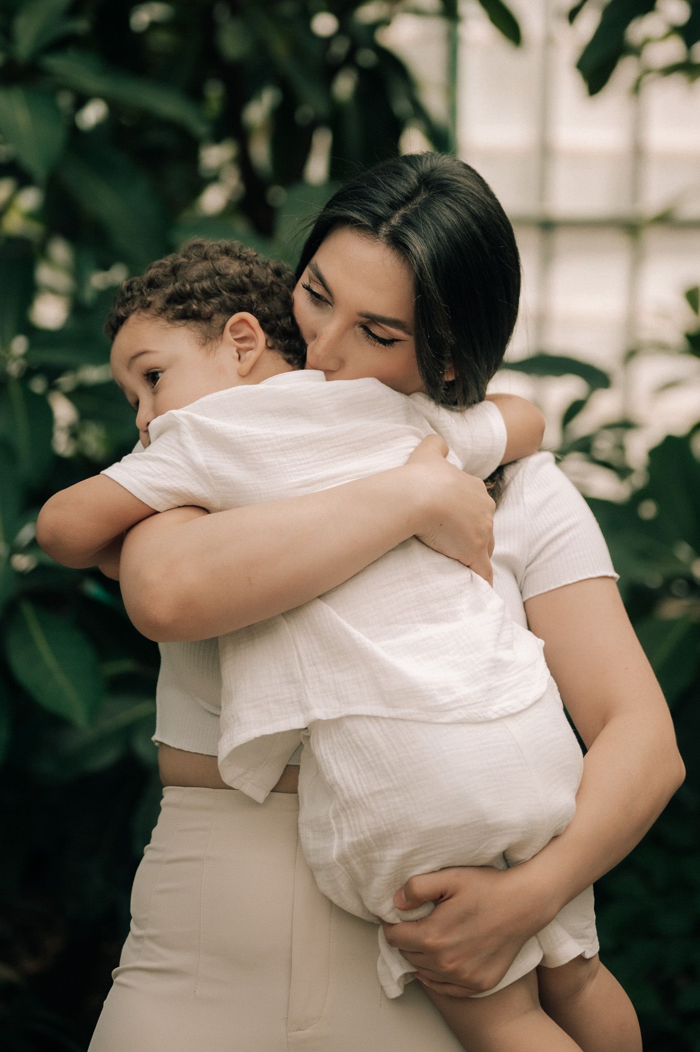 A mother lovingly embraces her young child in a tender hug surrounded by green foliage.