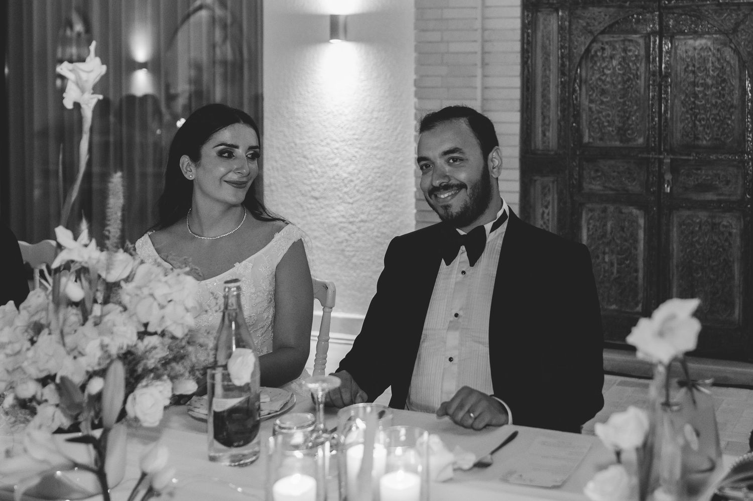 Black and white photo of smiling bride and groom seated at wedding reception table with candles and flowers.