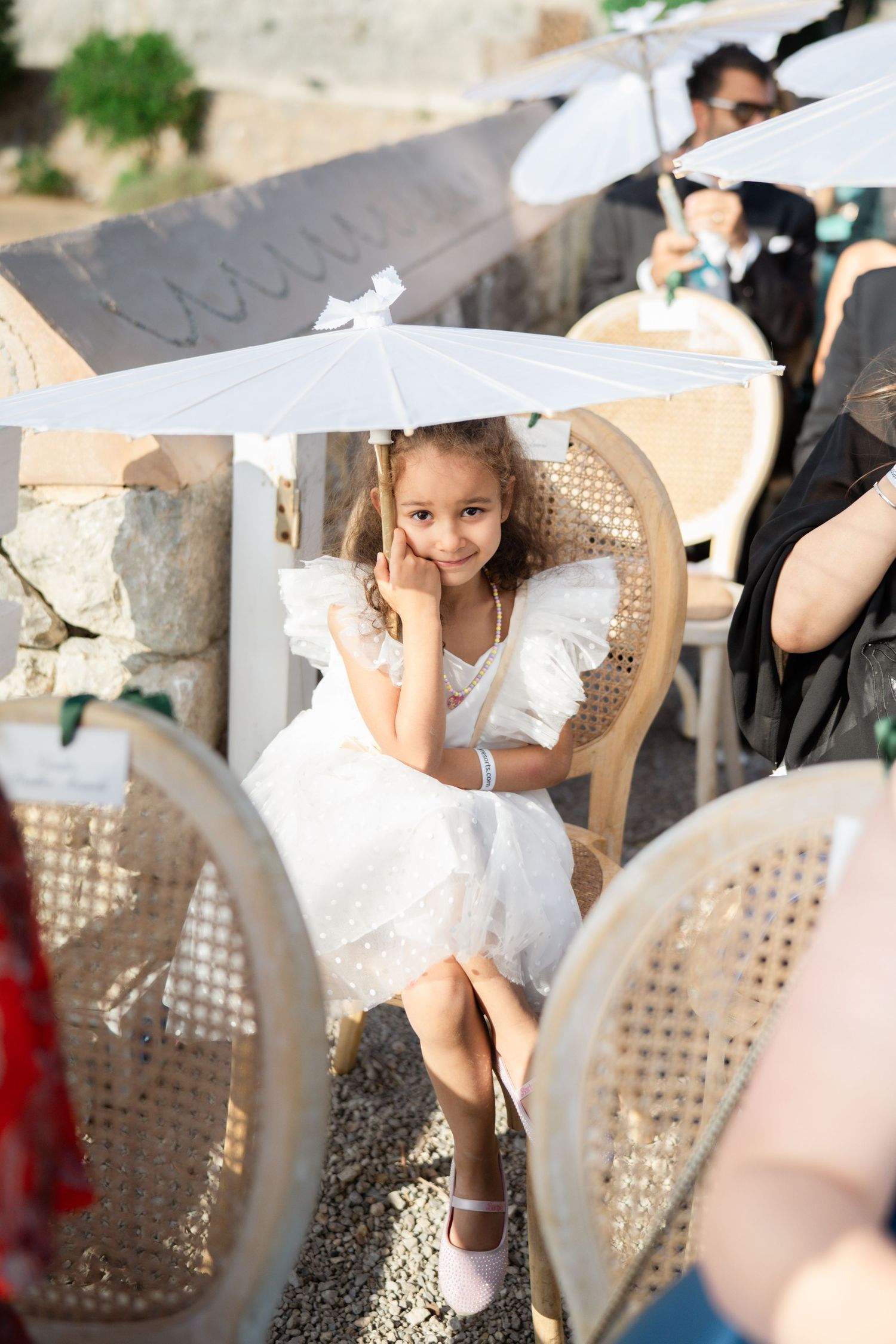 A young flower girl in a white dress sits under a parasol at an outdoor wedding ceremony.
