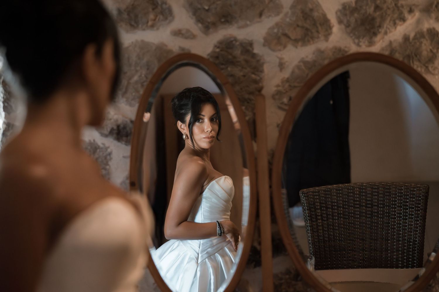 Elegant bride in strapless white gown gazing into oval mirror in rustic stone-walled room.