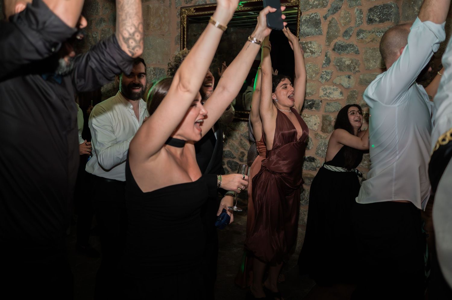 Guests dancing with arms raised at a lively wedding reception in a rustic stone venue.