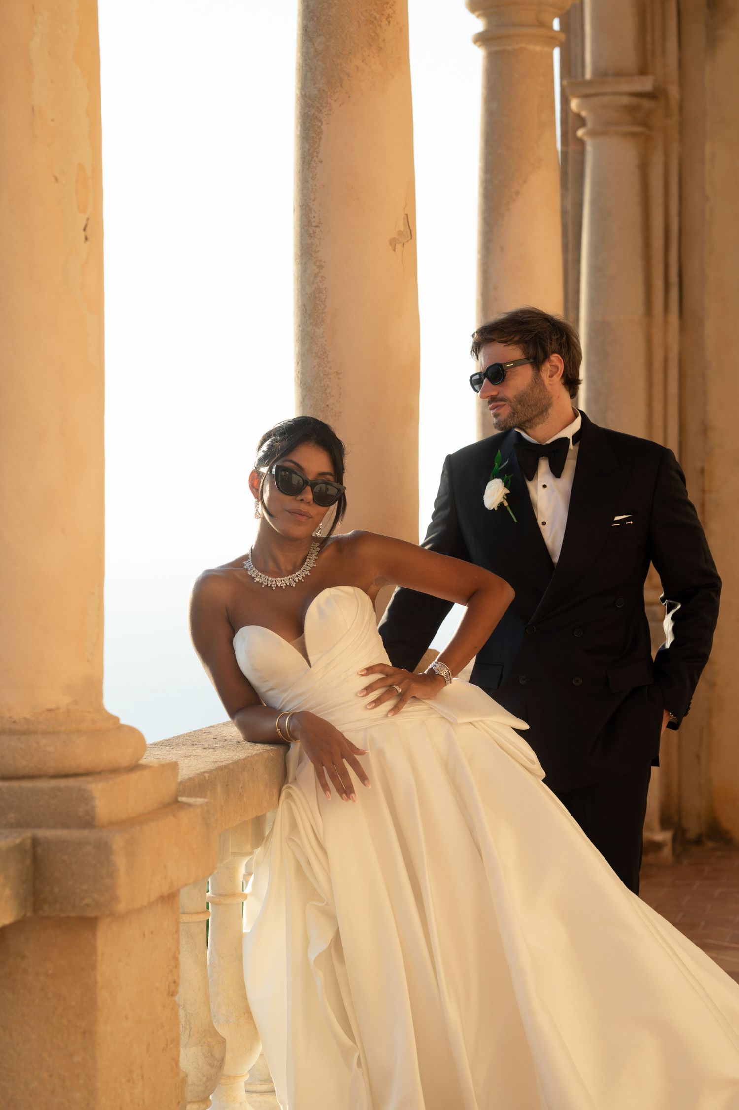 Stylish bride and groom pose in sunglasses between stone columns at Mediterranean wedding venue.