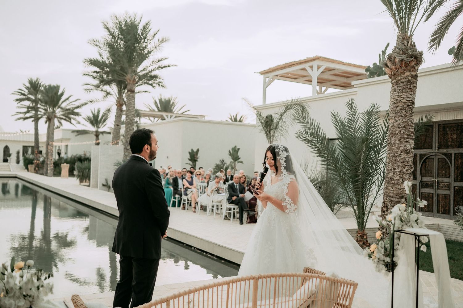 Bride in white ball gown walks toward groom at outdoor wedding ceremony beside reflecting pool with palm trees.