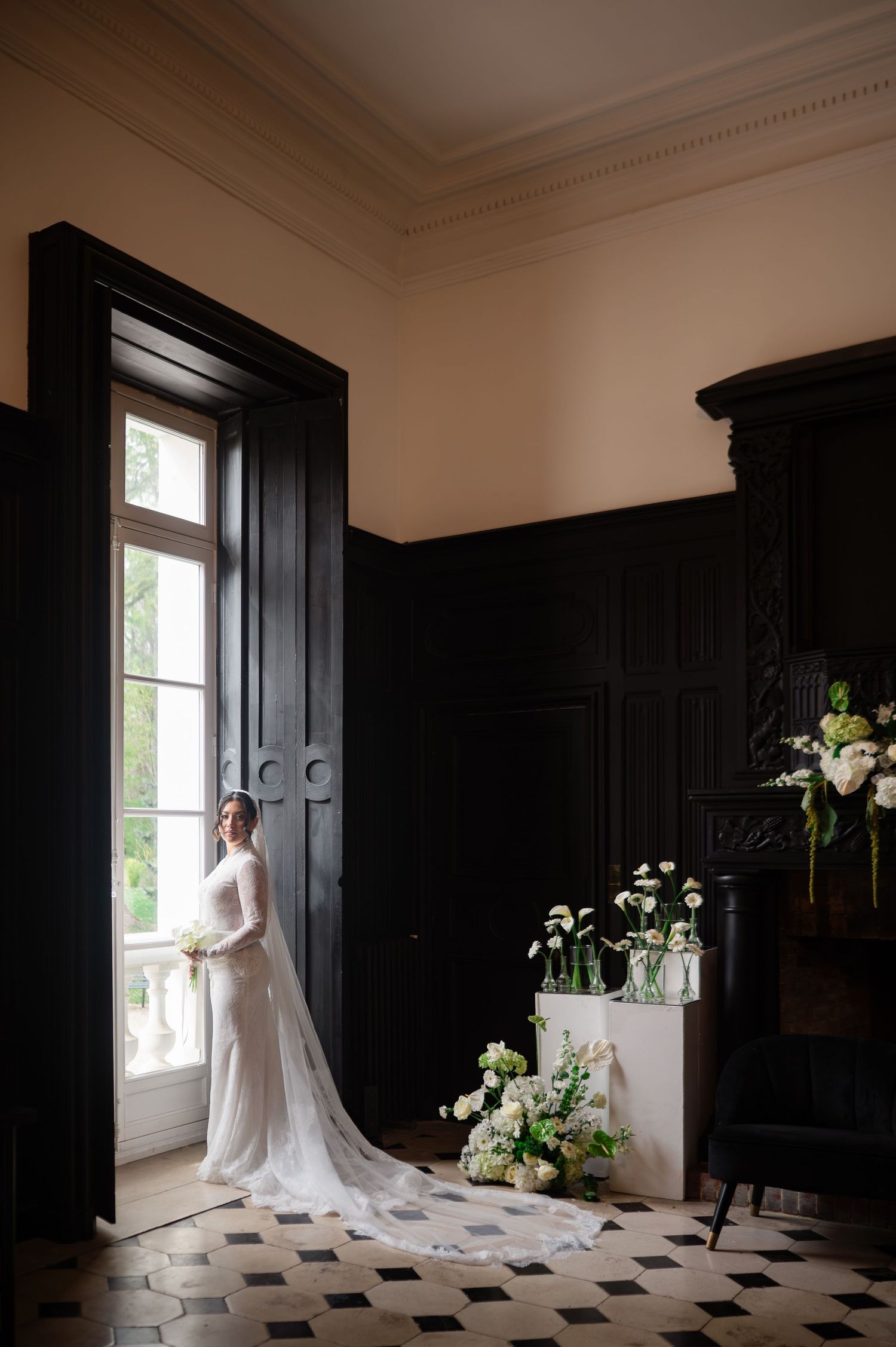 Bride in a long white gown with cathedral veil standing by black doors in a classic French château.