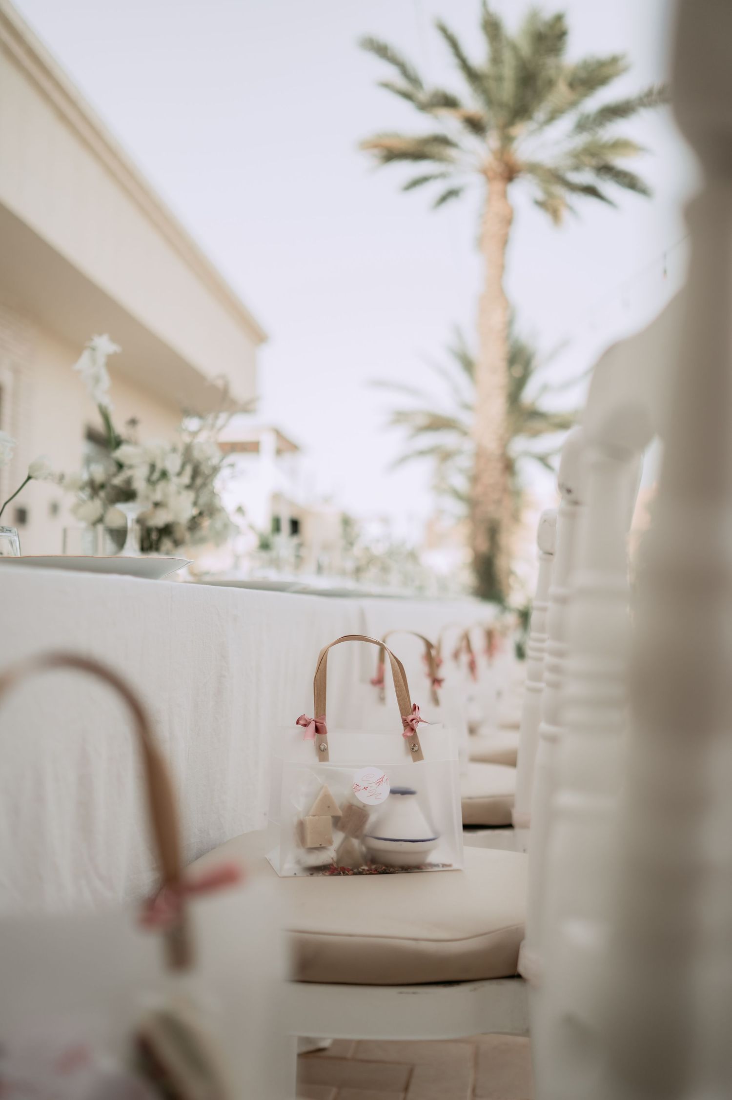 Elegant outdoor wedding reception table setting with white decor and favor bags under swaying palm trees.
