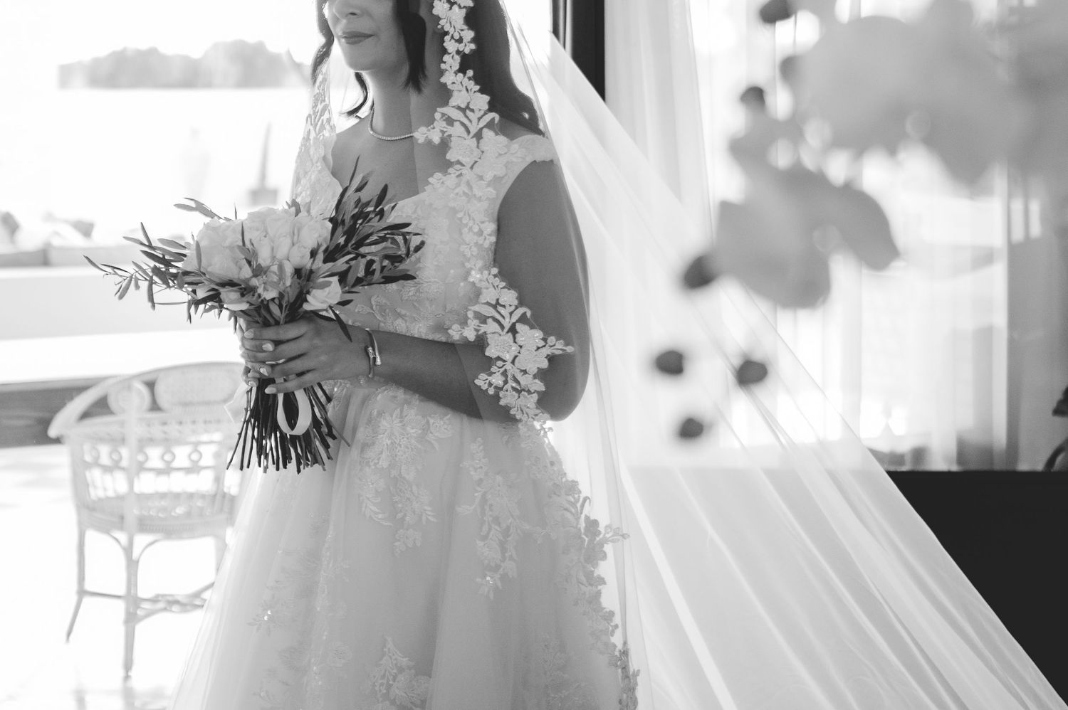 Black and white photo of bride in floral lace wedding dress holding bouquet by window.